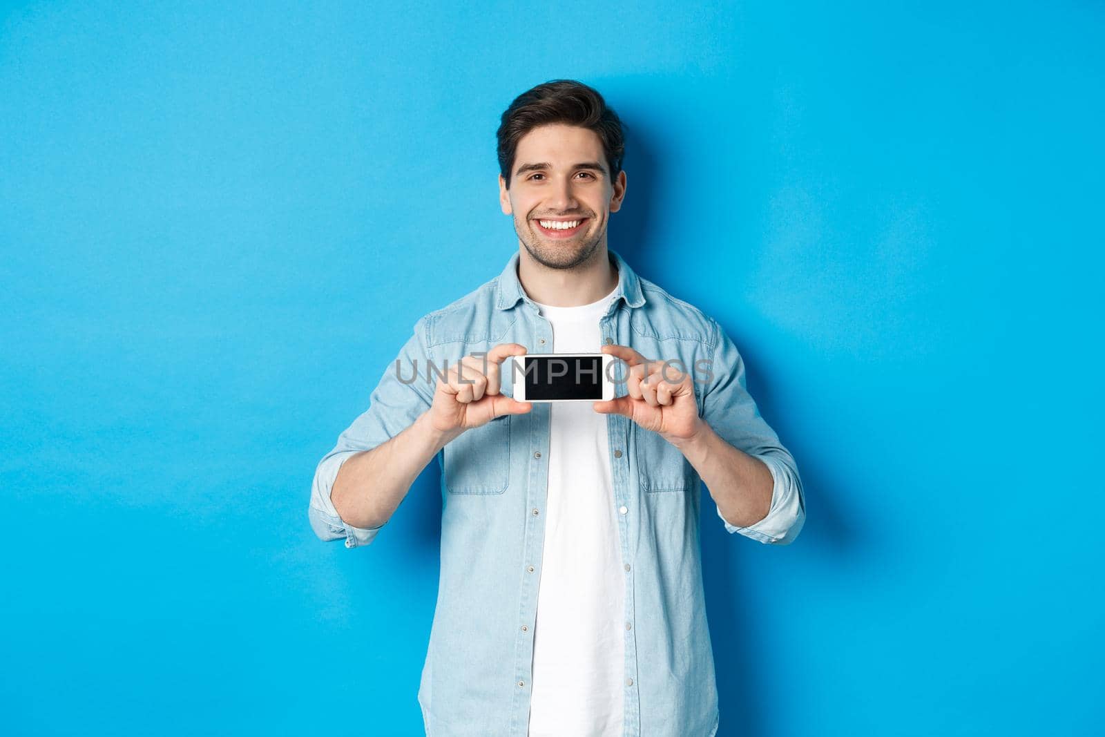 Handsome smiling man showing smartphone screen, standing against blue background for copy space.