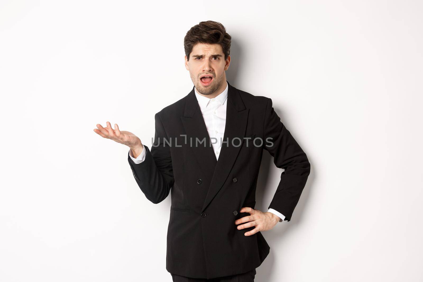 Portrait of arrogant man in black suit, looking confused and disappointed, complaining about something strange, standing over white background.