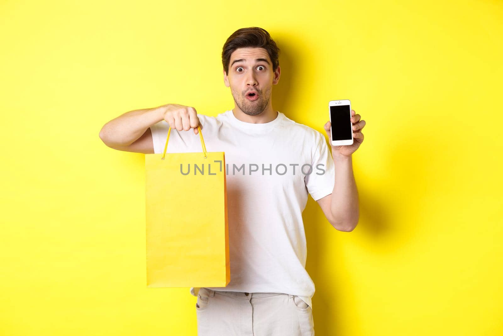 Surprised man holding shopping bag and showing smartphone screen, concept of mobile banking and app achievements, yellow background by Benzoix