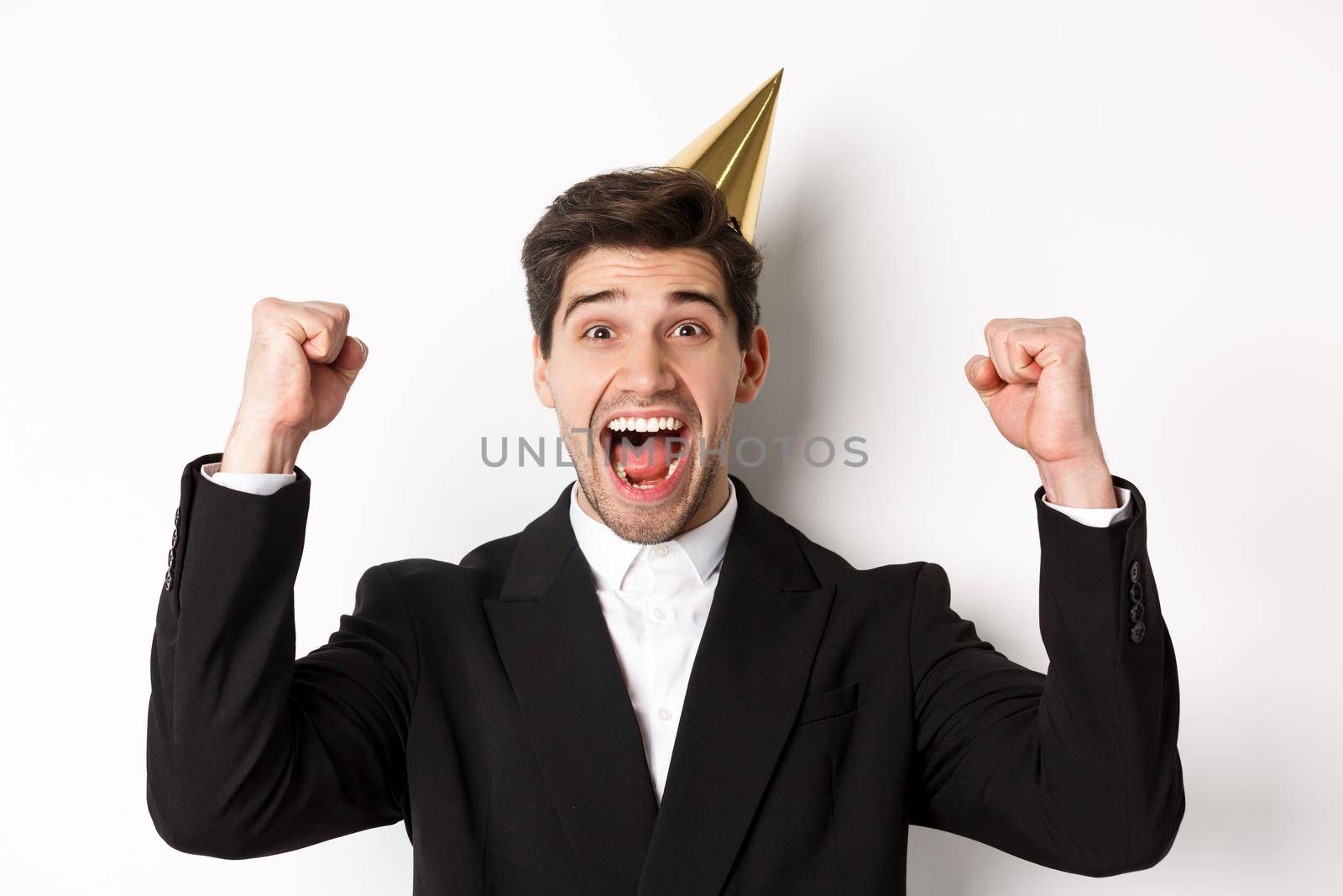 Close-up of happy good-looking man, wearing party hat and suit, raising hands up and rejoicing, celebrating new year, standing against white background by Benzoix