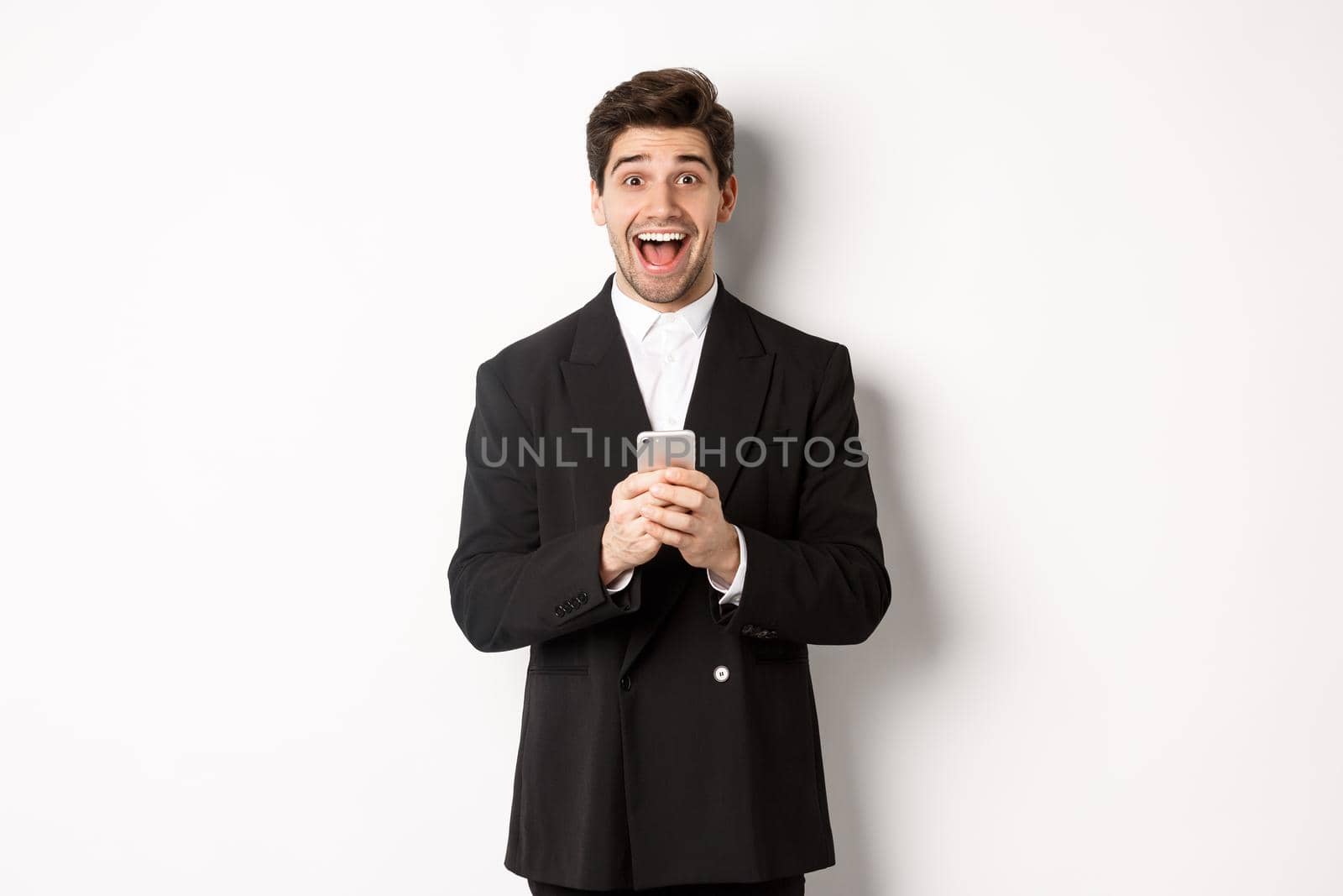 Image of excited handsome guy receiving promo offer on mobile phone, looking amazed and smiling, standing against white background by Benzoix