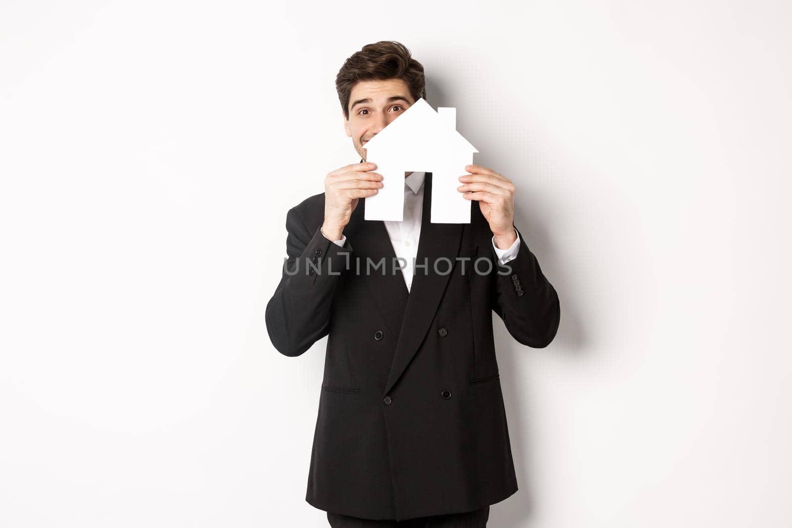 Image of handsome broker in black suit, showing house maket and smiling, selling homes, standing against white background by Benzoix