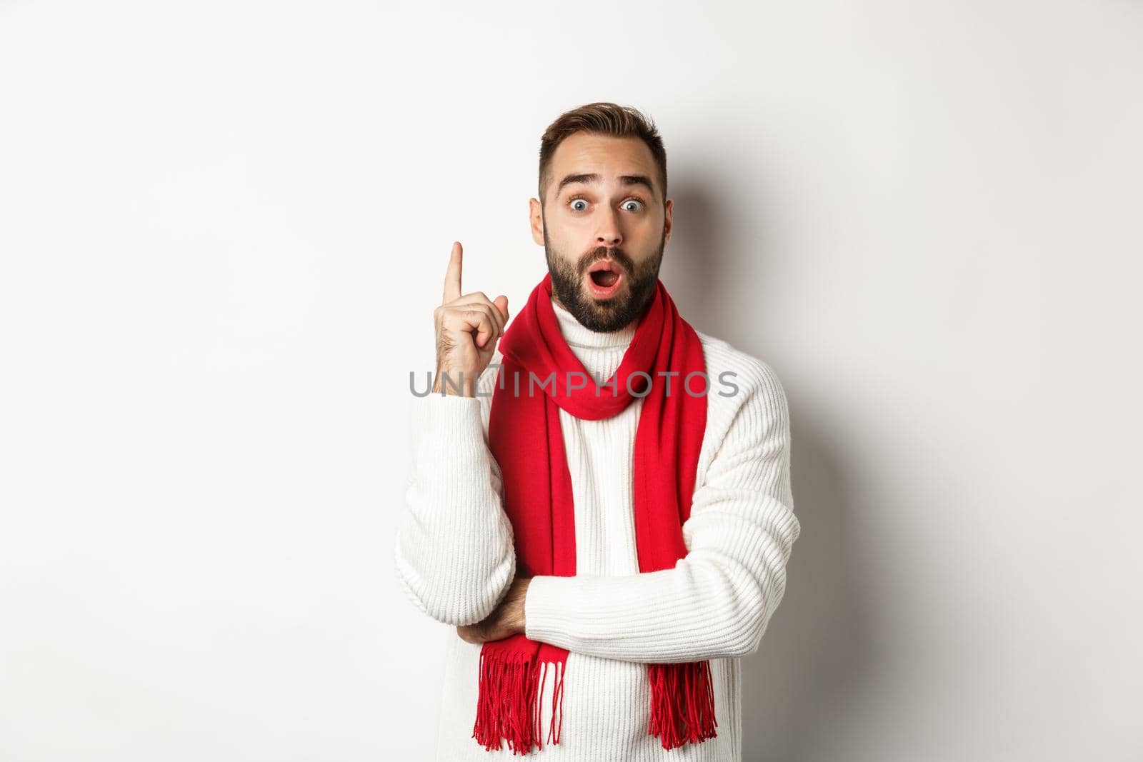 Christmas holidays and celebration concept. Excited bearded man having an idea, raising finger and suggesting plan, standing in red scarf with sweater, white background.