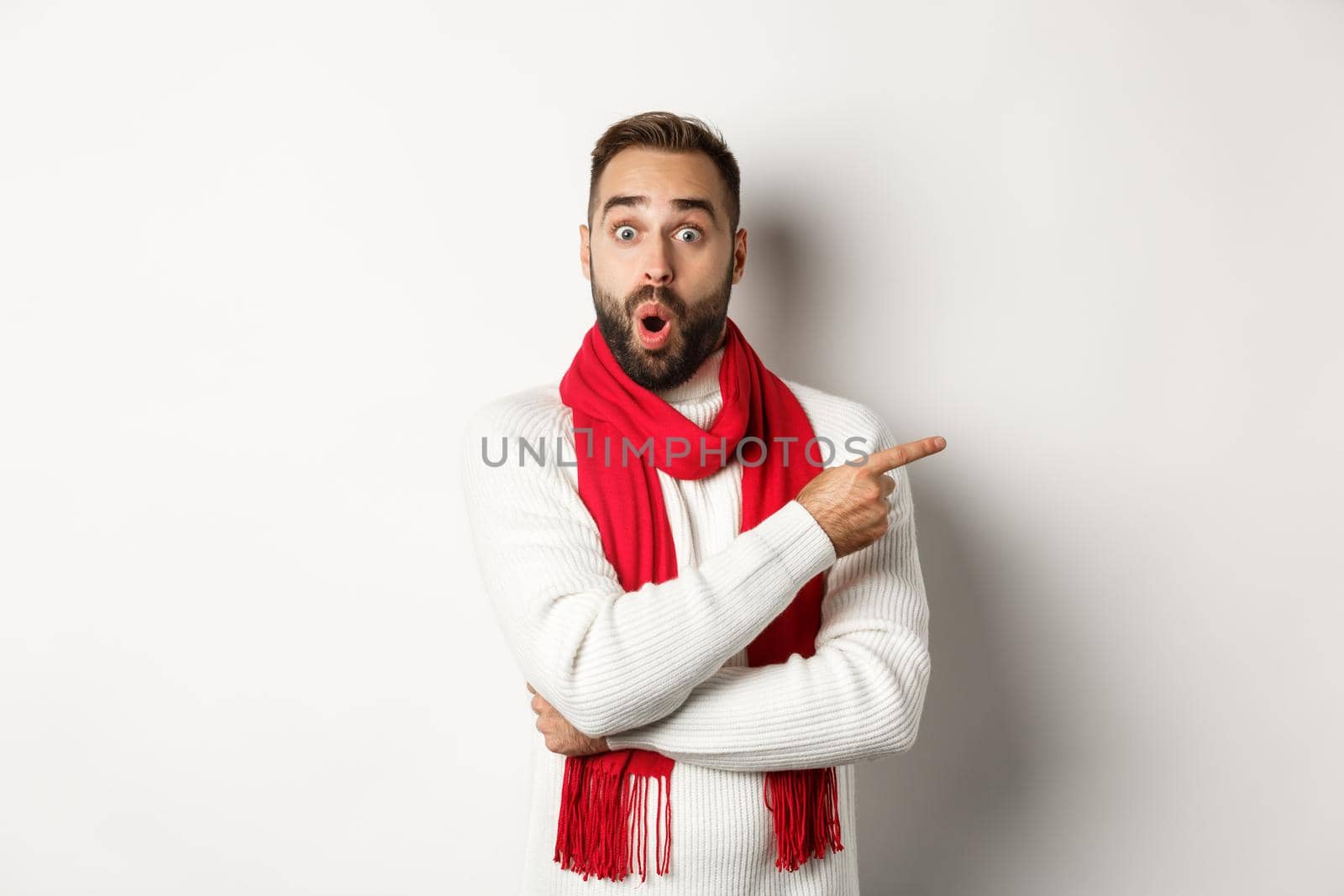 Christmas holidays and celebration concept. Surprised bearded man pointing finger right and looking impressed at camera, standing in red scarf and sweater, white background.