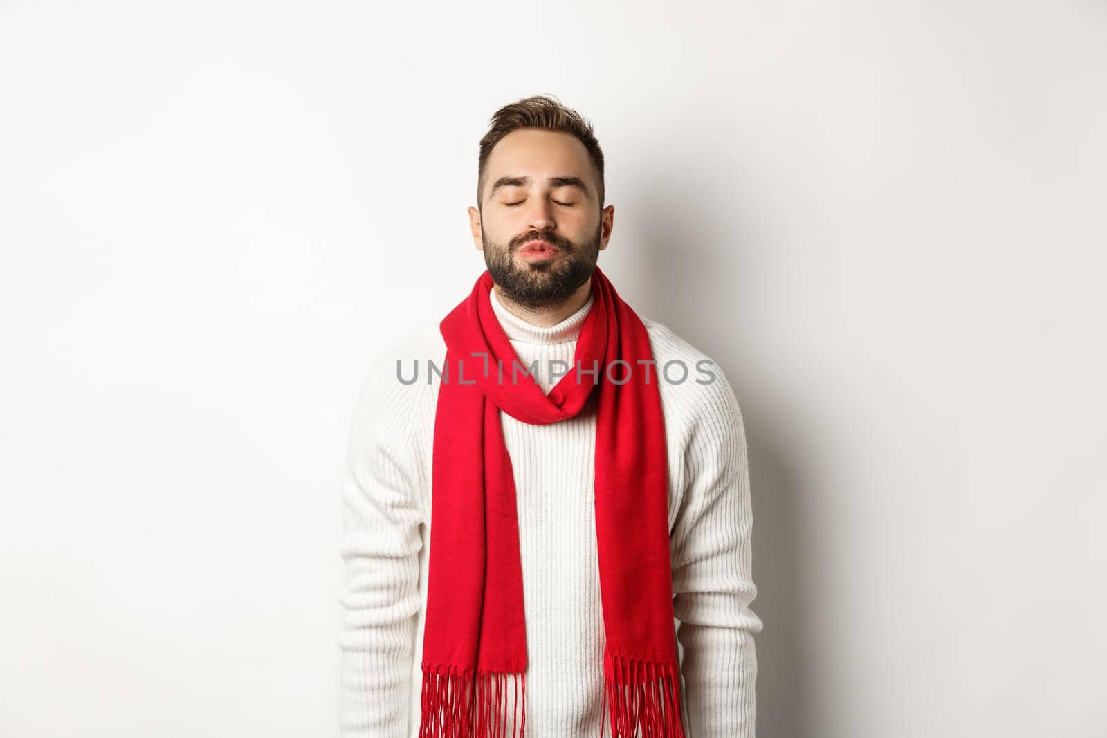 Handsome man standing with closed eyes and puckered lips, waiting for christmas kiss under mistletoe, standing over white background in winter scarf and sweater.