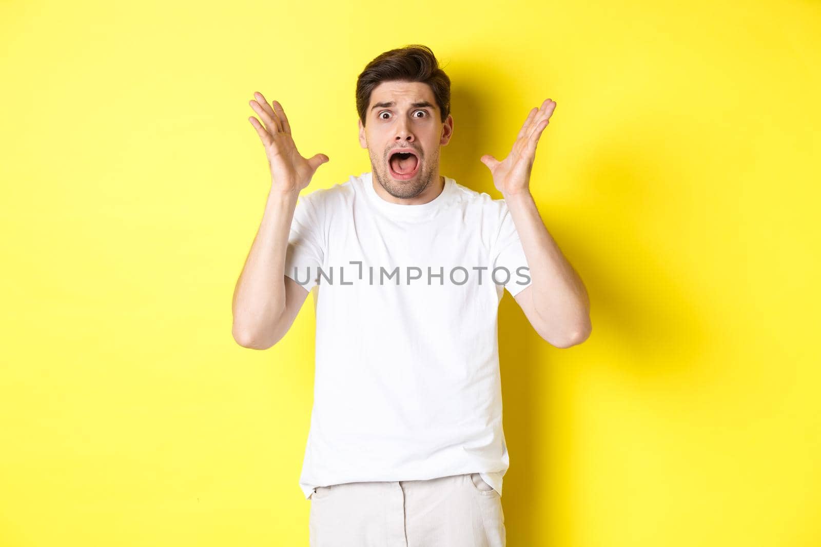 Frustrated and shocked guy panicking, screaming and looking scared, standing in white t-shirt over yellow background.
