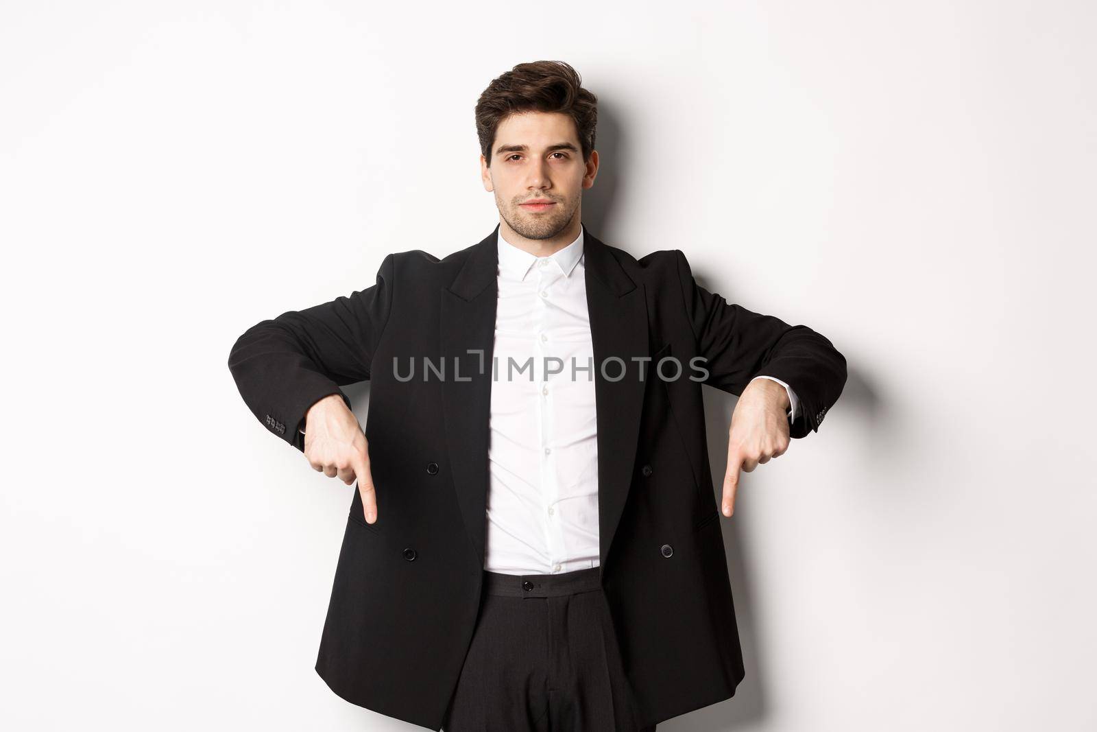 Image of handsome man dressed for formal party, wearing suit and pointing fingers down, showing advertisement or making announcement, standing over white background.
