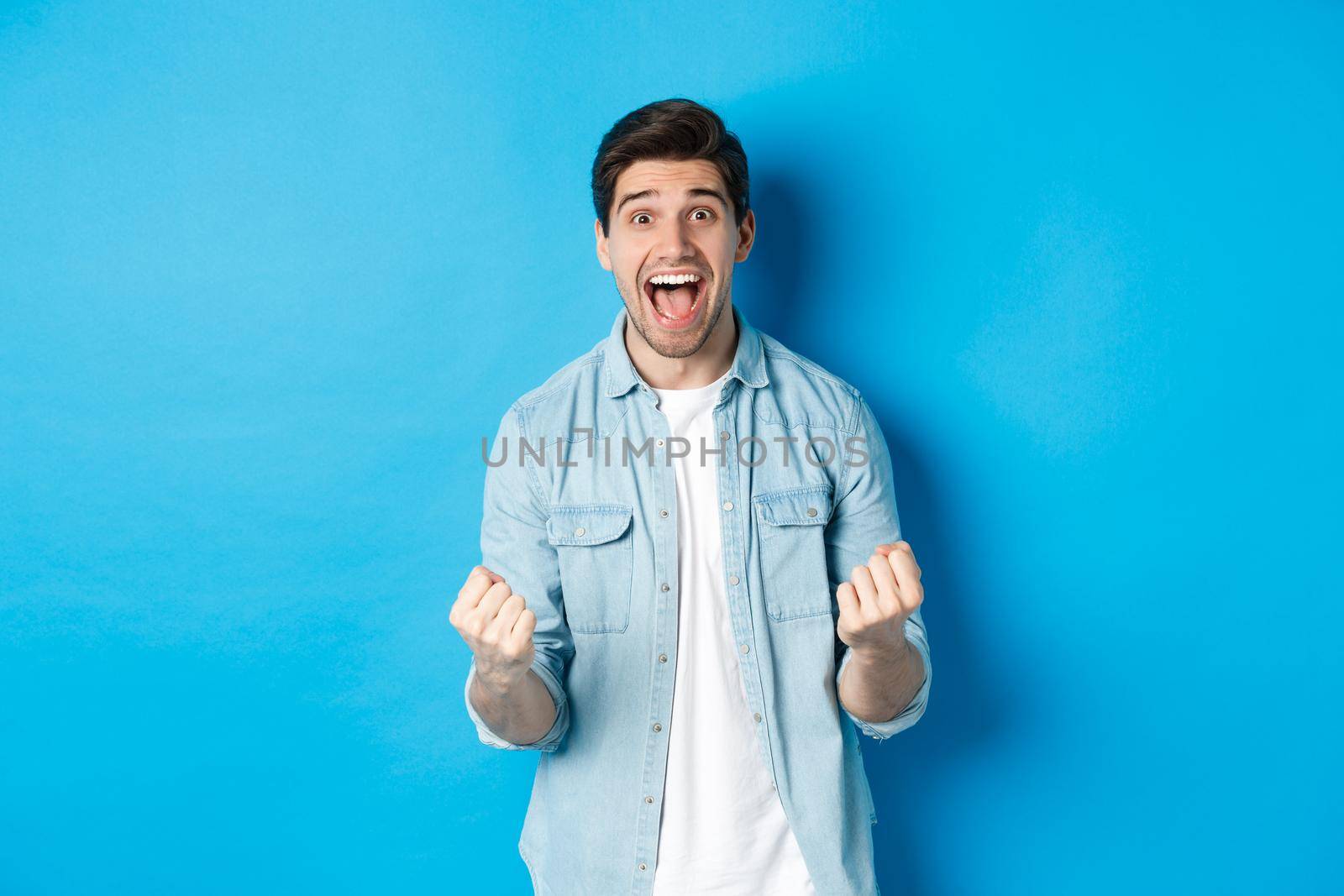 Excited handsome man winning, feeling lucky and rejoicing, celebrating victory and shouting yes, standing against blue background.
