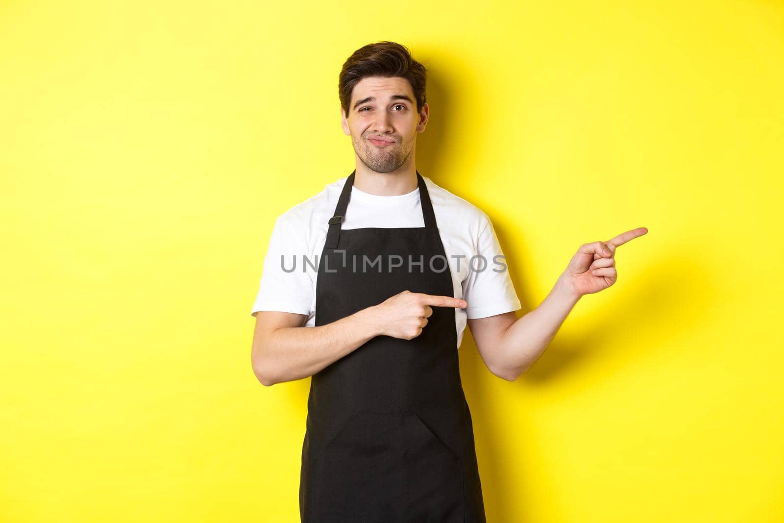 Doubtful barista in black apron pointing fingers right, looking skeptical and unamused, standing over yellow background by Benzoix
