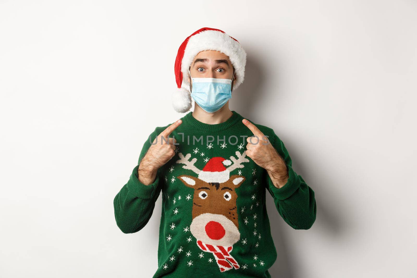 Christmas during pandemic, covid-19 concept. Man in in Santa hat pointing at his face mask, celebrating New Year and social distancing, white background.