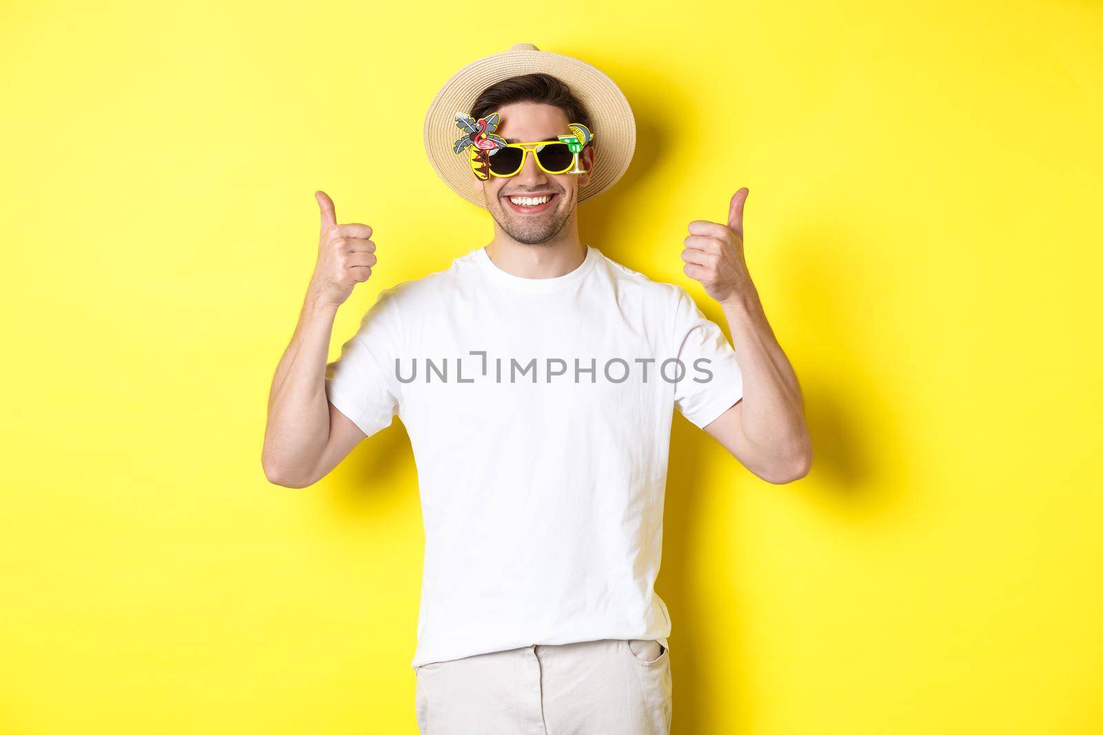 Concept of tourism and lifestyle. Image of smiling tourist showing thumbs-up, enjoying trip and recommending, wearing summer hat and sunglasses, yellow background.
