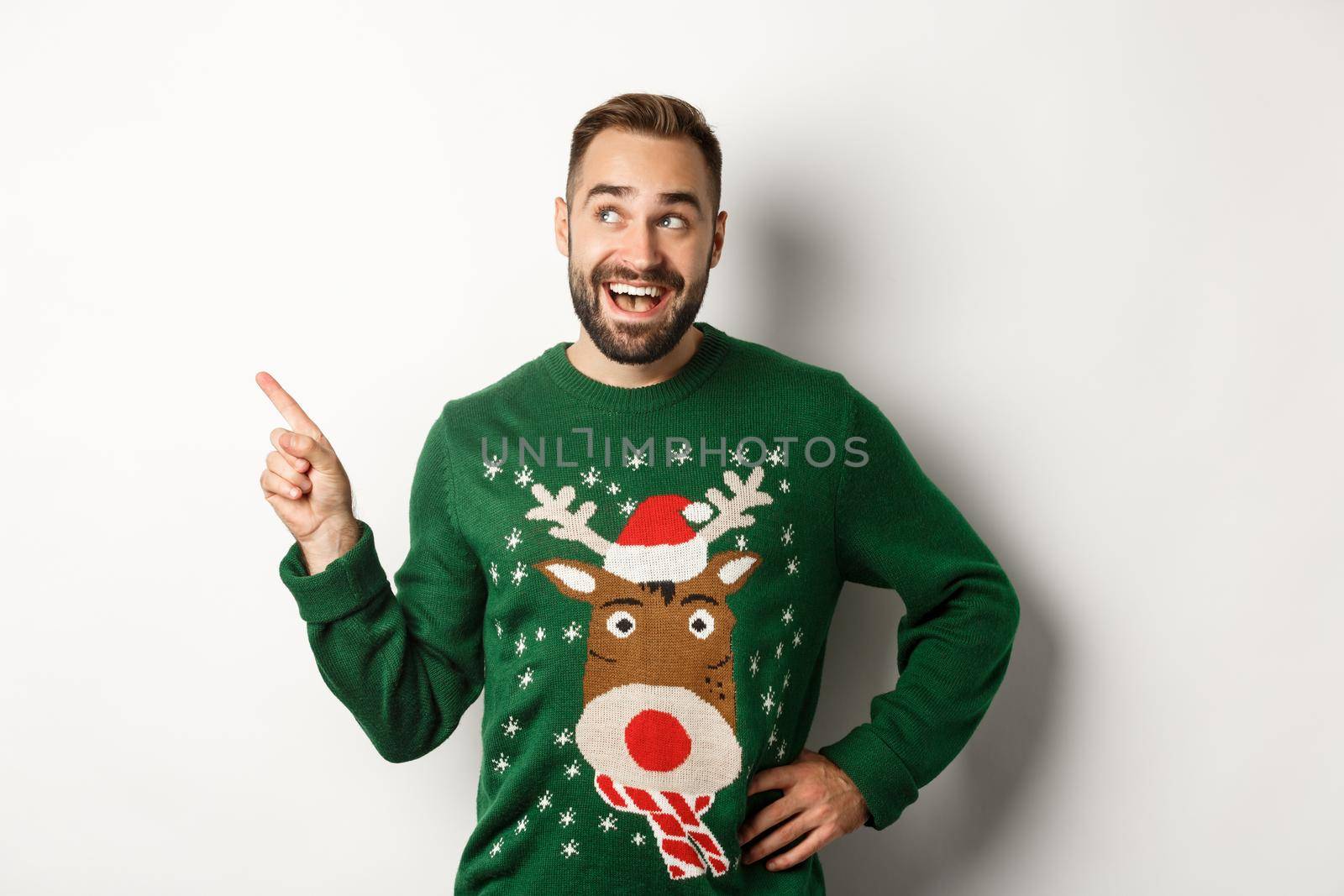 New year celebration and winter holidays concept. Dreamy bearded man in green christmas sweater, pointing at upper left corner and smiling amused, white background.