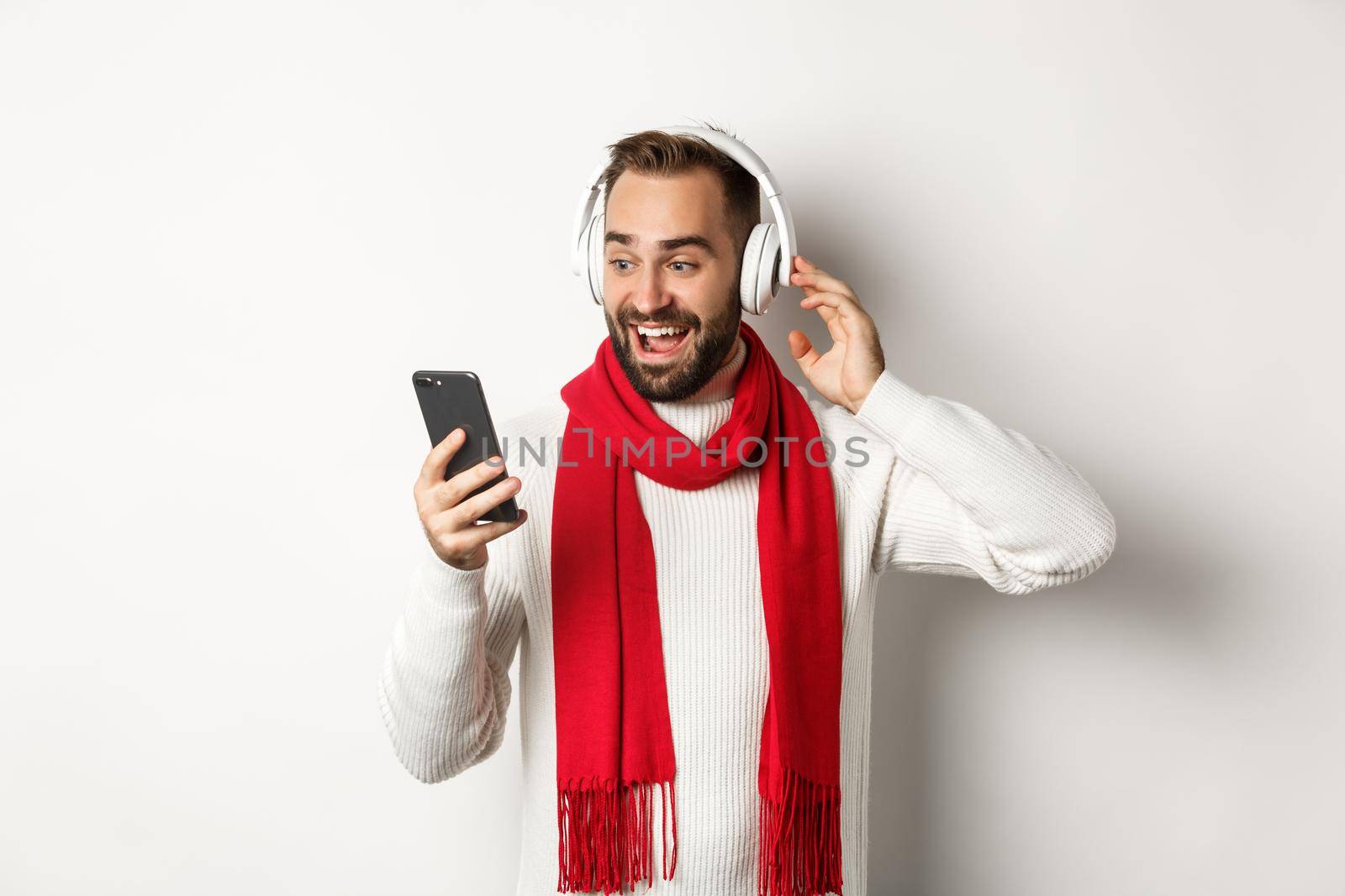 Winter holidays and technology concept. Happy man listening music in headphones, looking amazed at mobile screen, standing over white background.