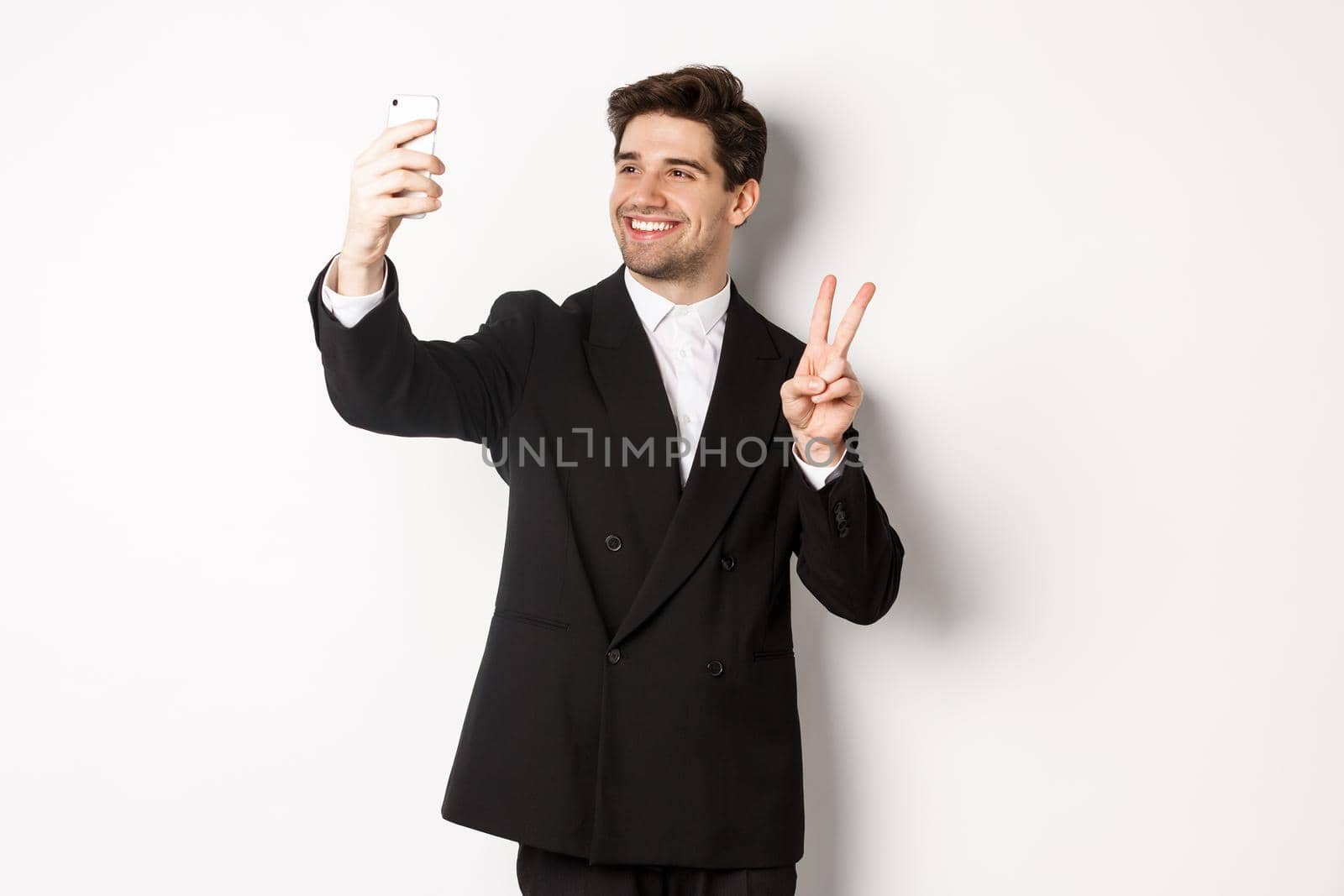 Portrait of good-looking man taking selfie on new year party, wearing suit, taking photo on smartphone and showing peace sign, standing against white background by Benzoix