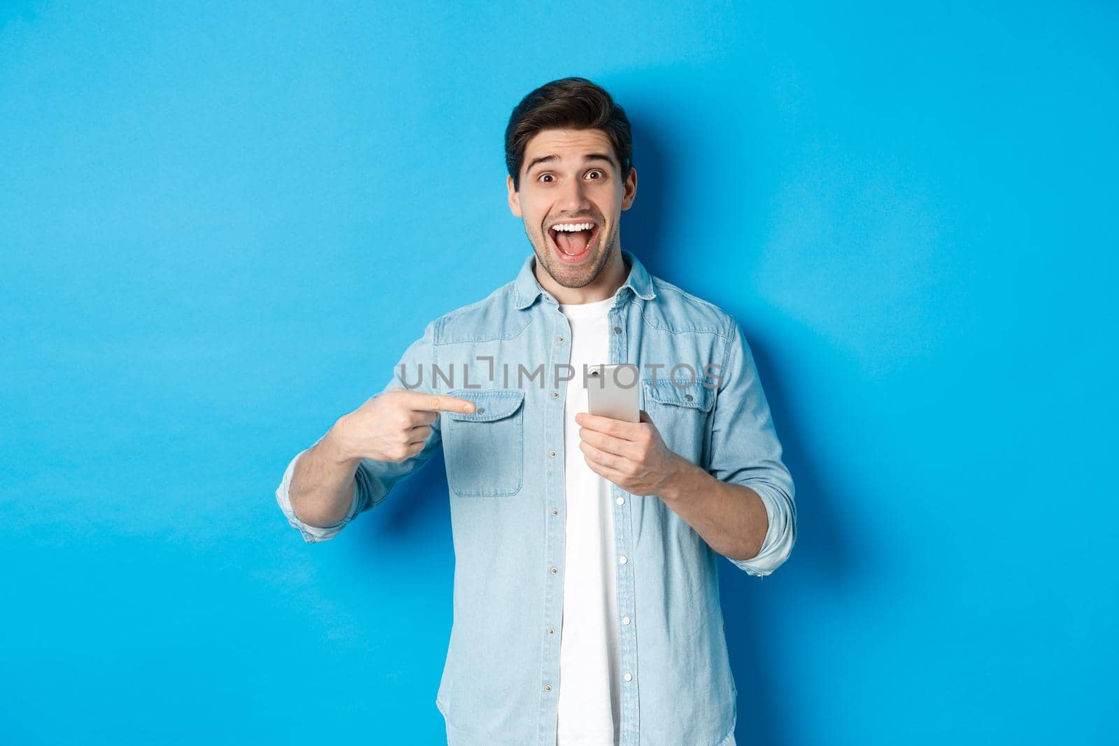 Concept of online shopping, applications and technology. Happy smiling man pointing finger at phone, standing amazed against blue background by Benzoix