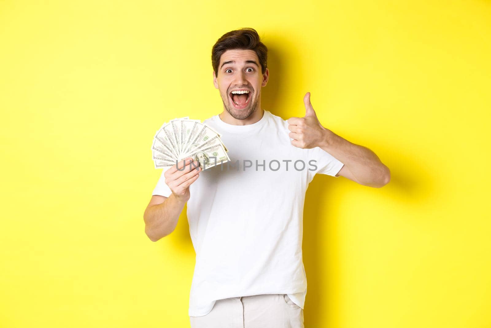Excited man holding money, showing thumb-up in approval, got credit or loan, standing over yellow background by Benzoix