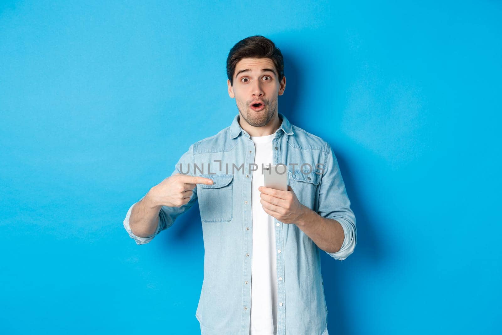 Concept of online shopping, applications and technology. Impressed man pointing finger at mobile phone and looking amazed, recommending app, standing over blue background by Benzoix