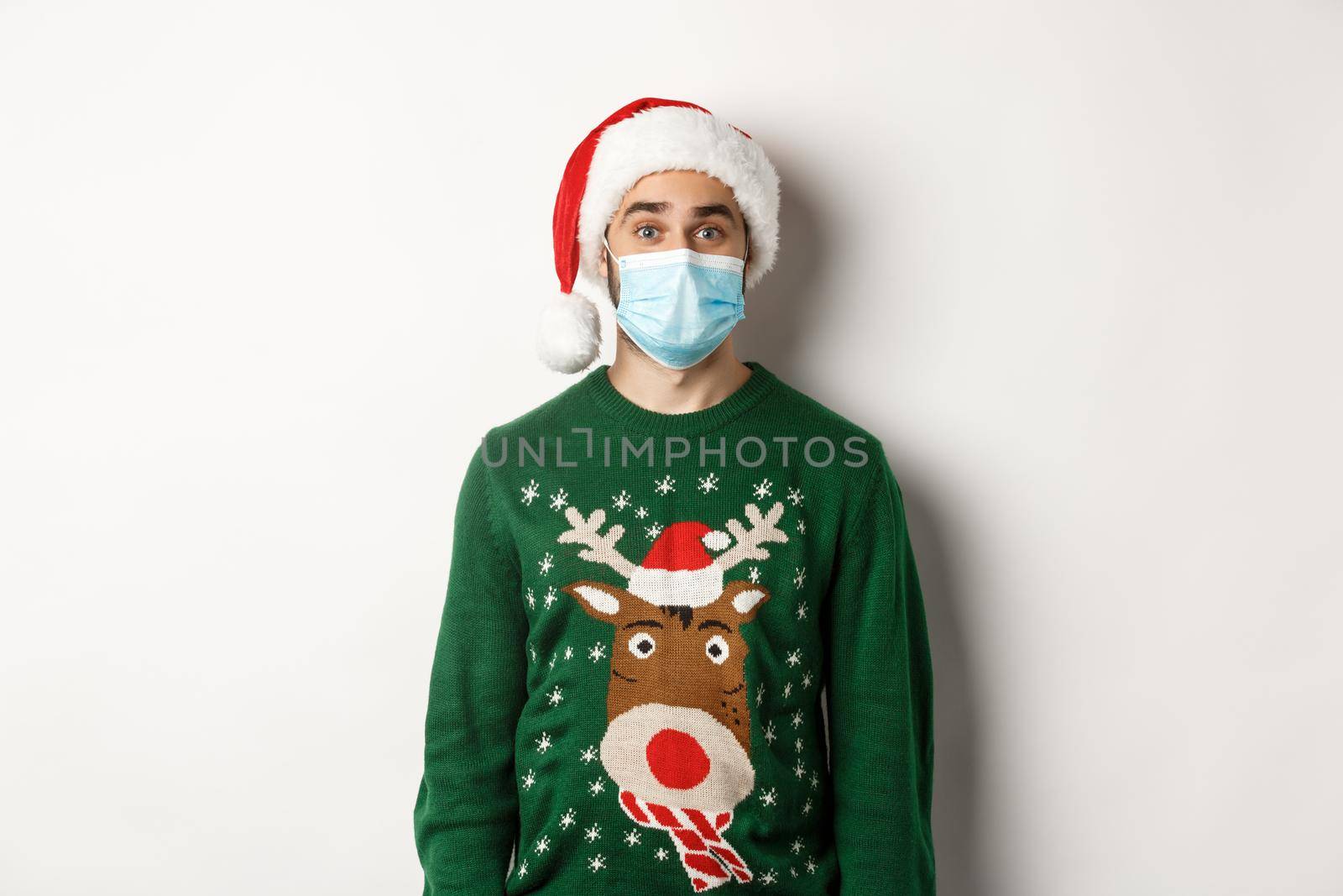 Christmas during pandemic, covid-19 concept. Young man in Santa hat and face mask celebrating New Year party with preventive measures, white background.