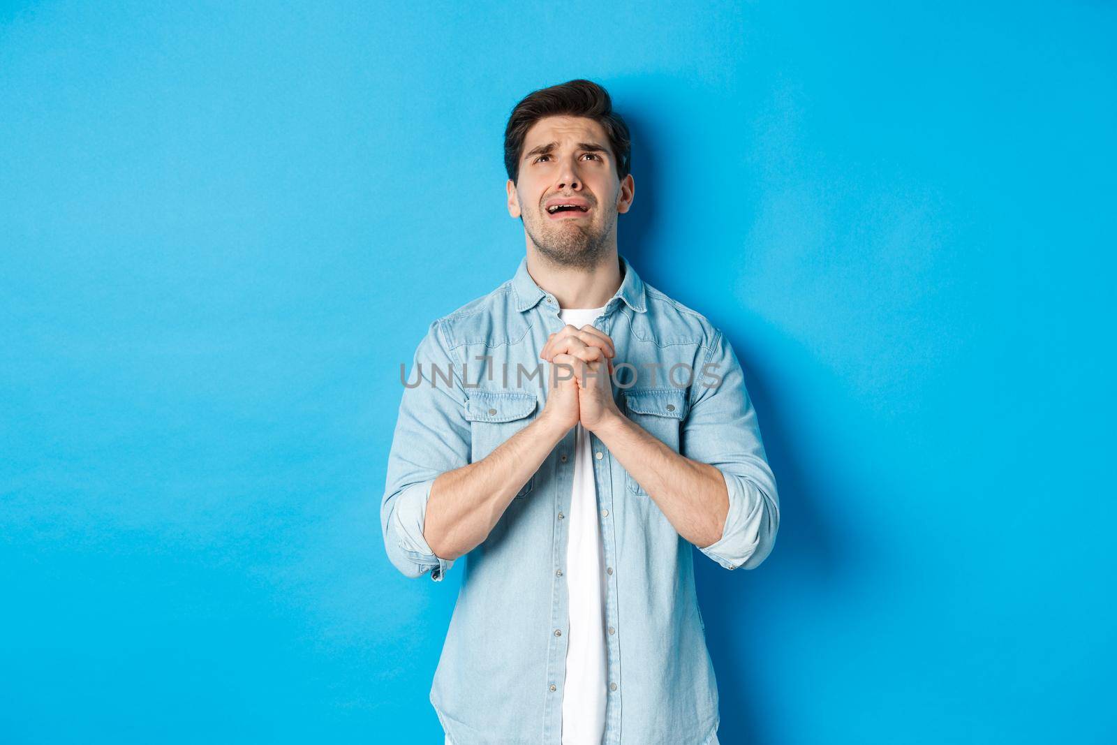Desperate man begging and crying for help, pleading to god, looking up and holding hands in pray, standing against blue background.