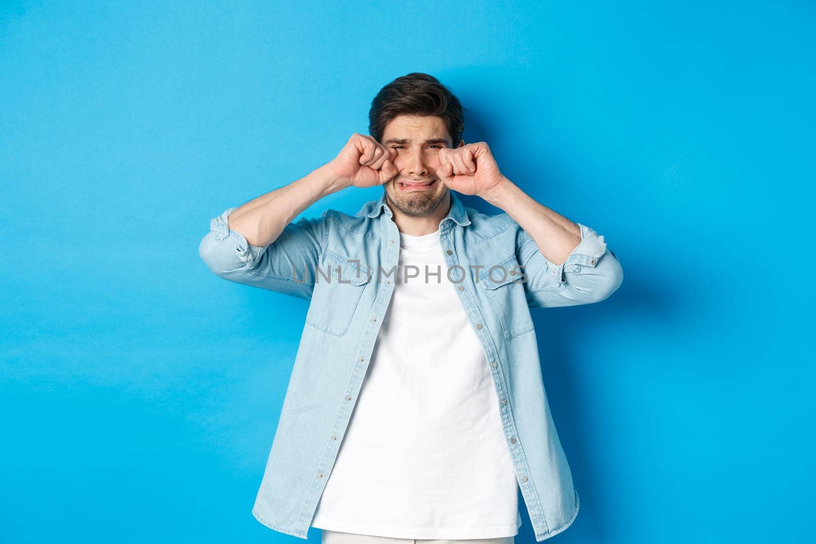 Sad timid man crying, wiping tears off face and sobbing, looking offended and upset, standing over blue background by Benzoix