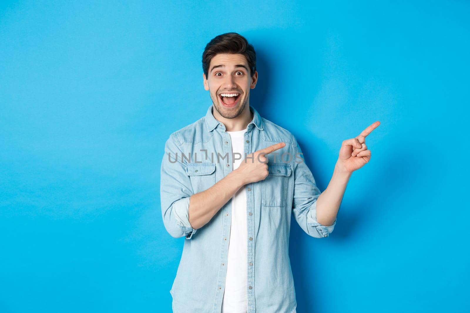 Image of excited handsome man in casual outfit, showing advertisement, pointing fingers right at copy space and smiling, standing against blue background by Benzoix