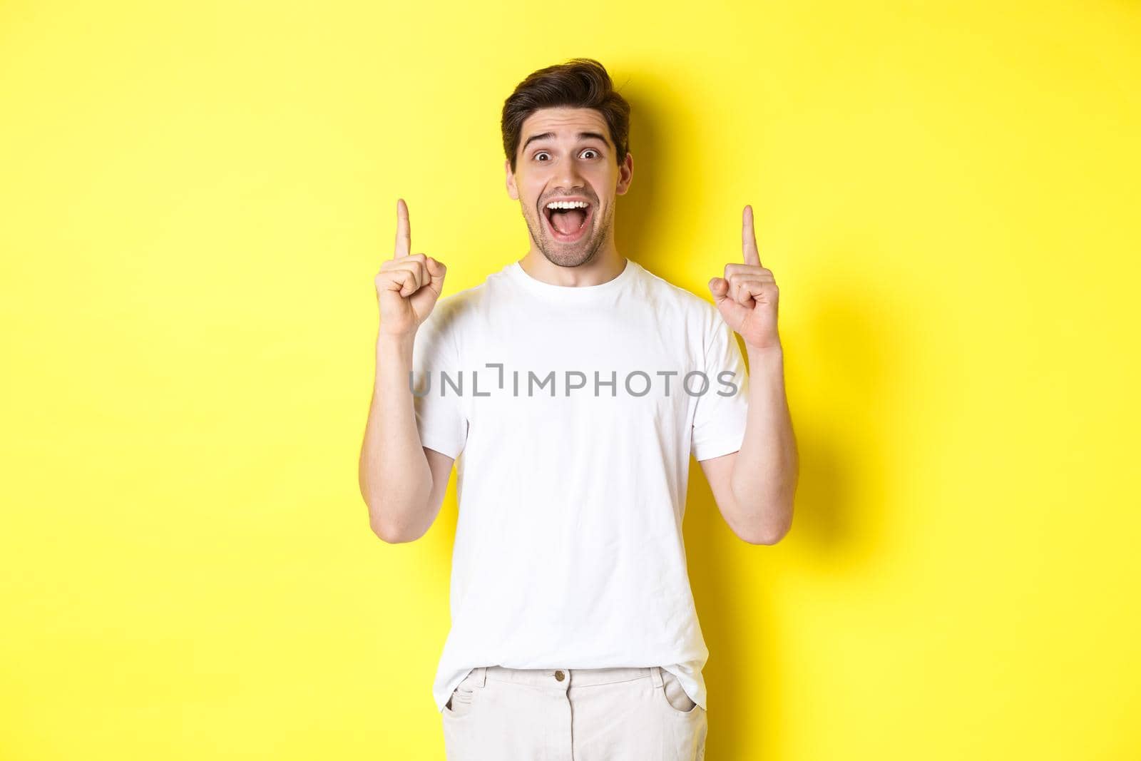 Portrait of excited handsome man in white t-shirt, pointing fingers up, showing offer, standing against yellow background by Benzoix