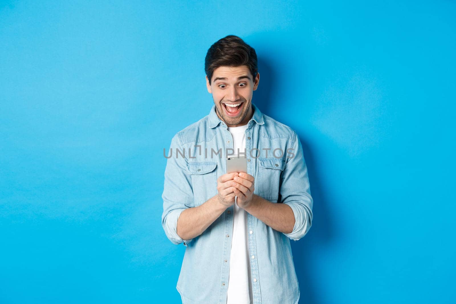 Image of excited man smiling while looking at mobile phone, shopping online on smartphone, standing against blue background.
