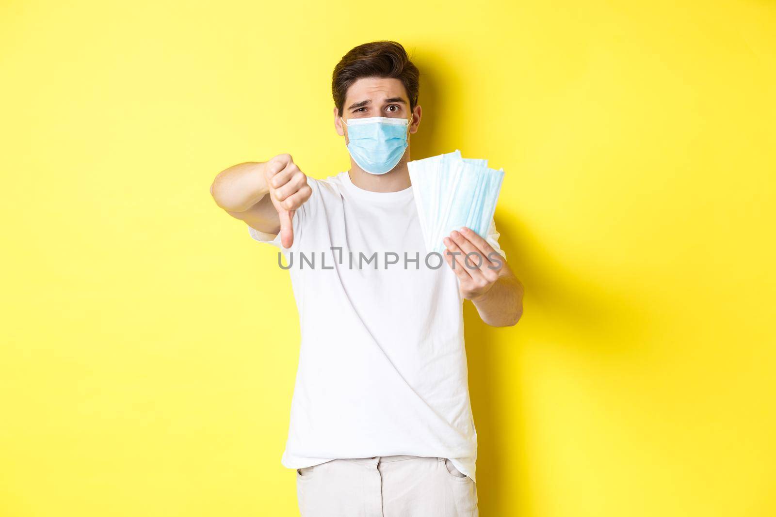 Concept of covid-19, quarantine and preventive measures. Man looking disappointed and showing thumb down, do not recommend bad medical masks, standing over yellow background.