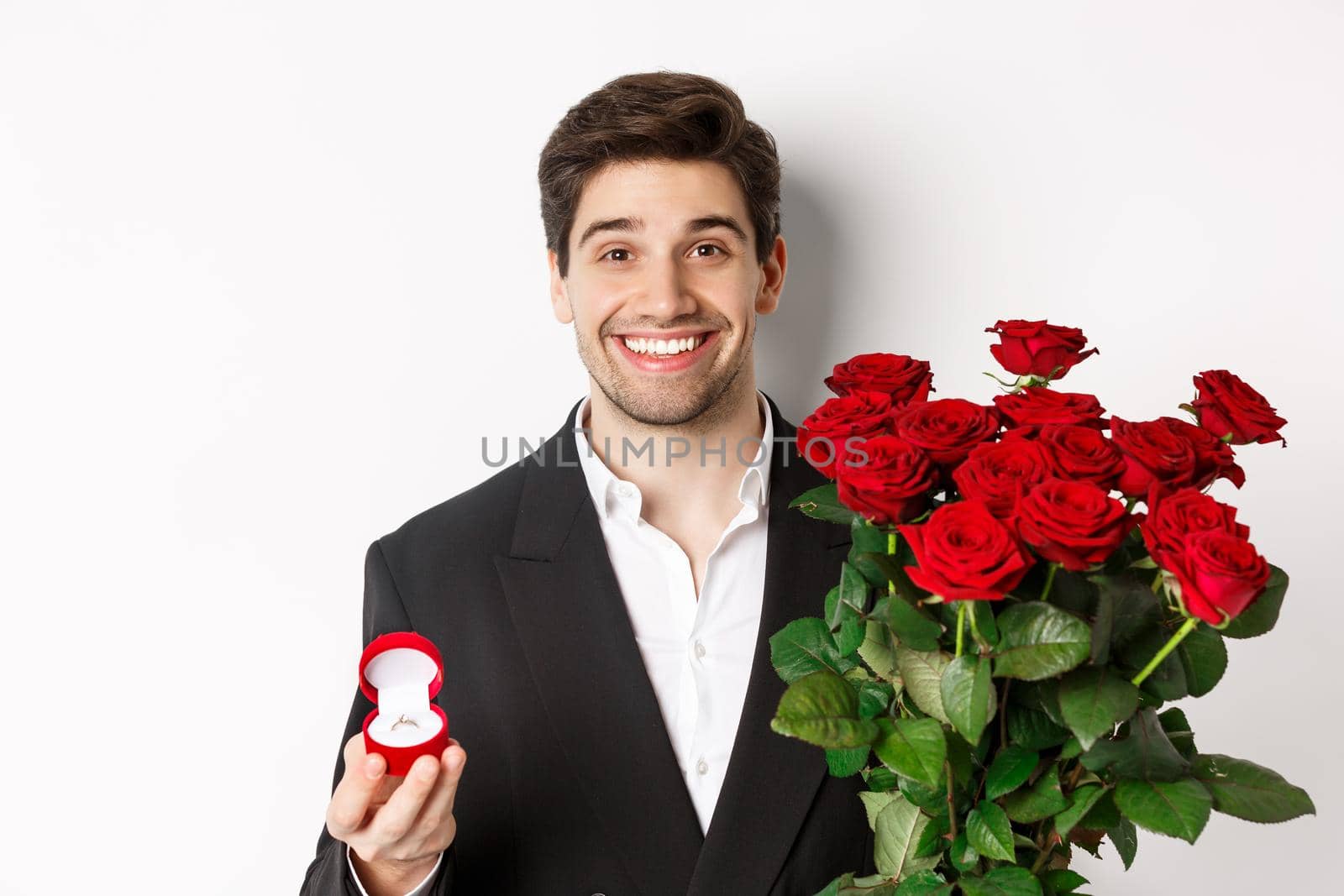 Close-up of attractive man in suit, holding bouquet of roses and engagement ring, making proposal, standing against white background by Benzoix