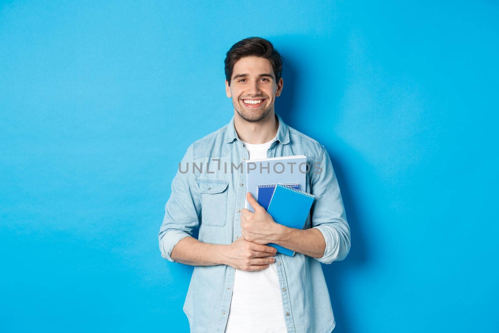 Young man holding notebooks and study material, smiling happy, standing over blue background.