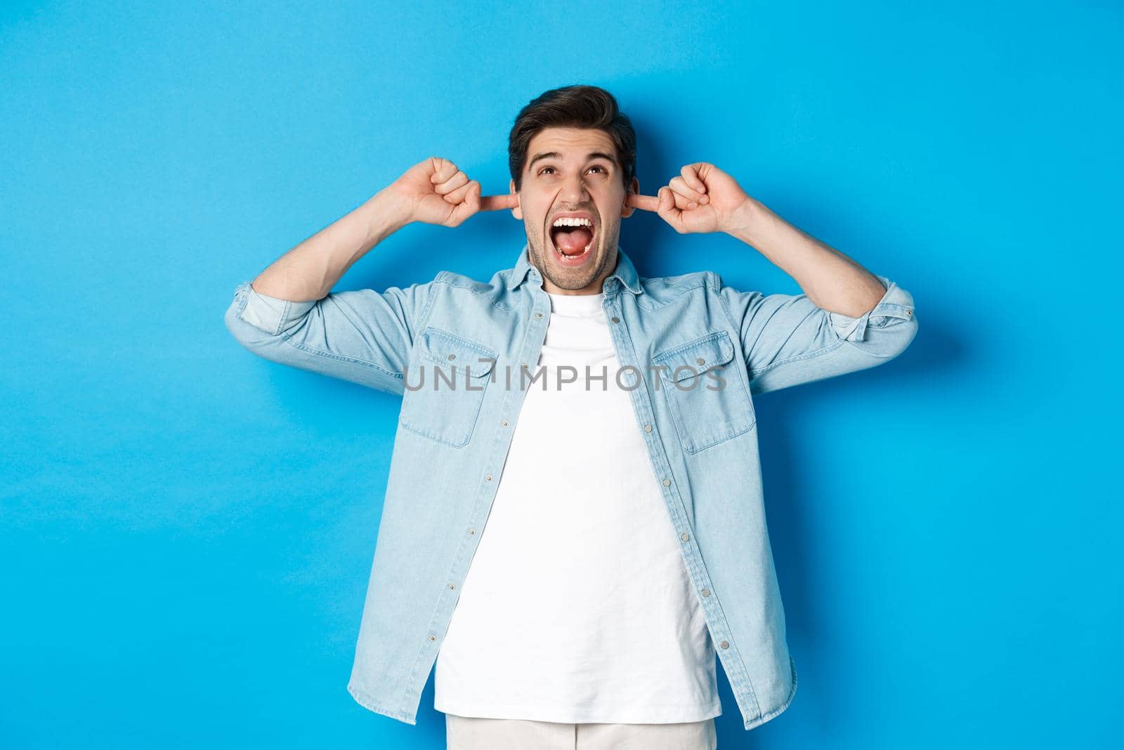 Irritated man close ears from noisy neighbours, looking up and shouting annoyed, hearing loud music, standing over blue background by Benzoix