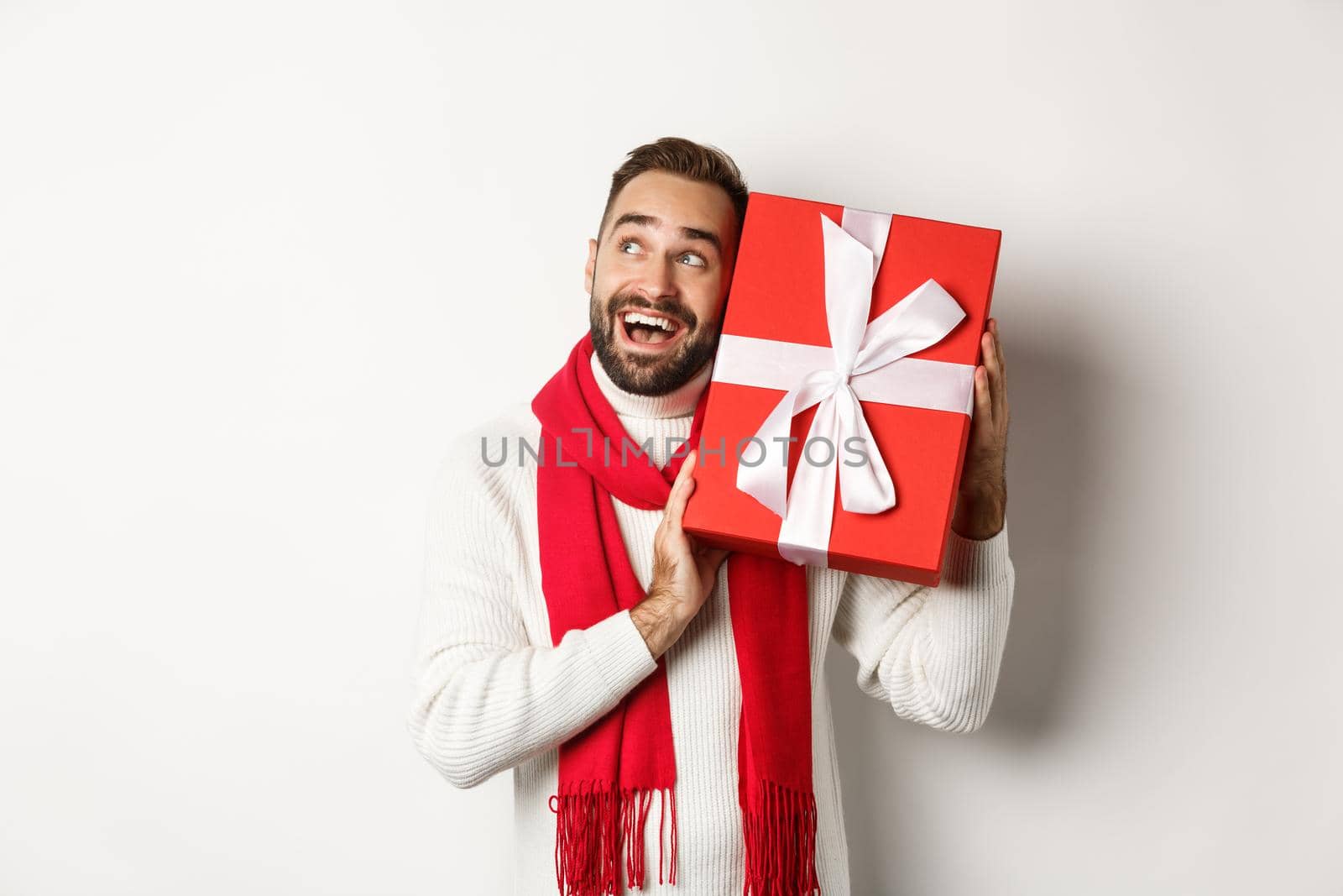 Concept of winter holidays. Happy man shaking gift box to guess what inside, looking curious, receive christmas presents, standing over white background.