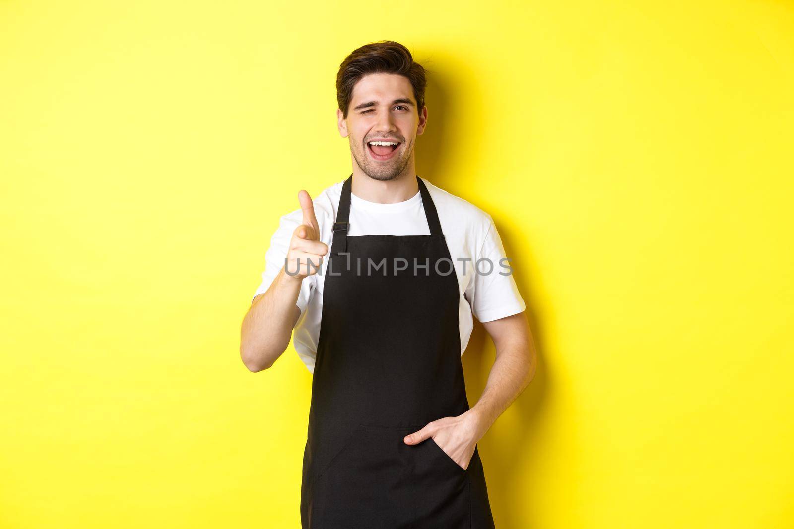 Handsome barista in black apron welcome guests to coffee shop, pointing finger gun and winking, standing against yellow background.