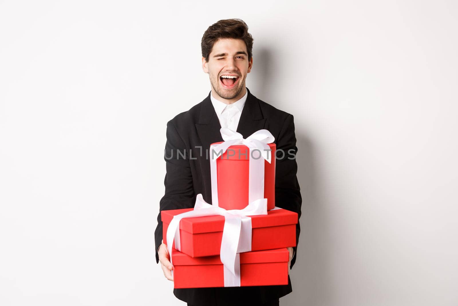 Image of handsome guy in black suit, holding gifts and winking at camera, have christmas presents, standing against white background.