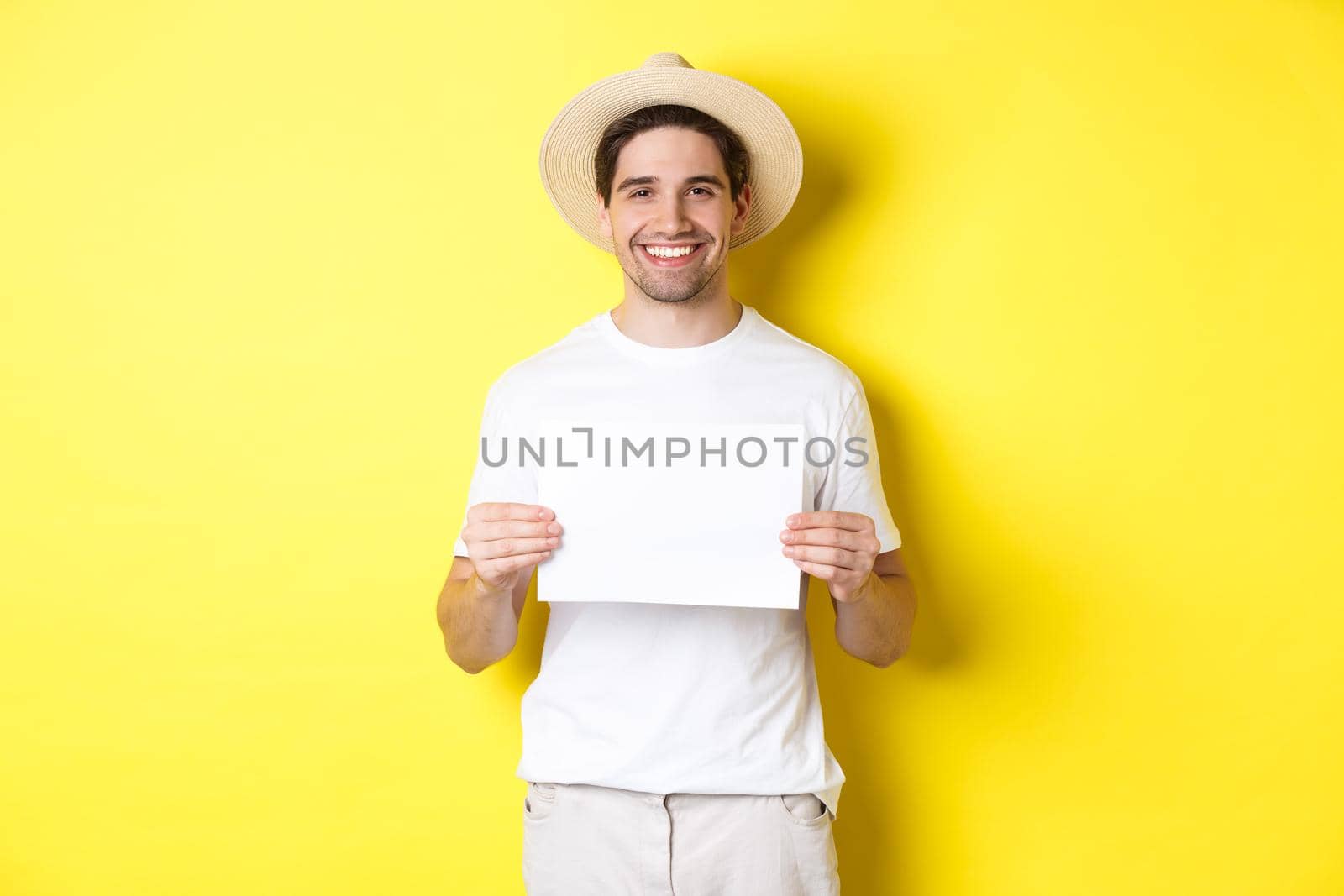Handsome young male tourist in summer hat smiling, holding blank piece of paper for your sign, standing over yellow background by Benzoix