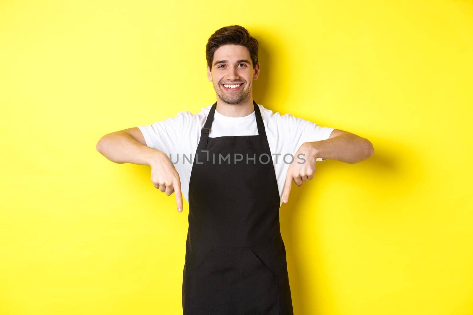 Friendly barista in black apron pointing fingers down, showing your logo banner, standing over yellow background.