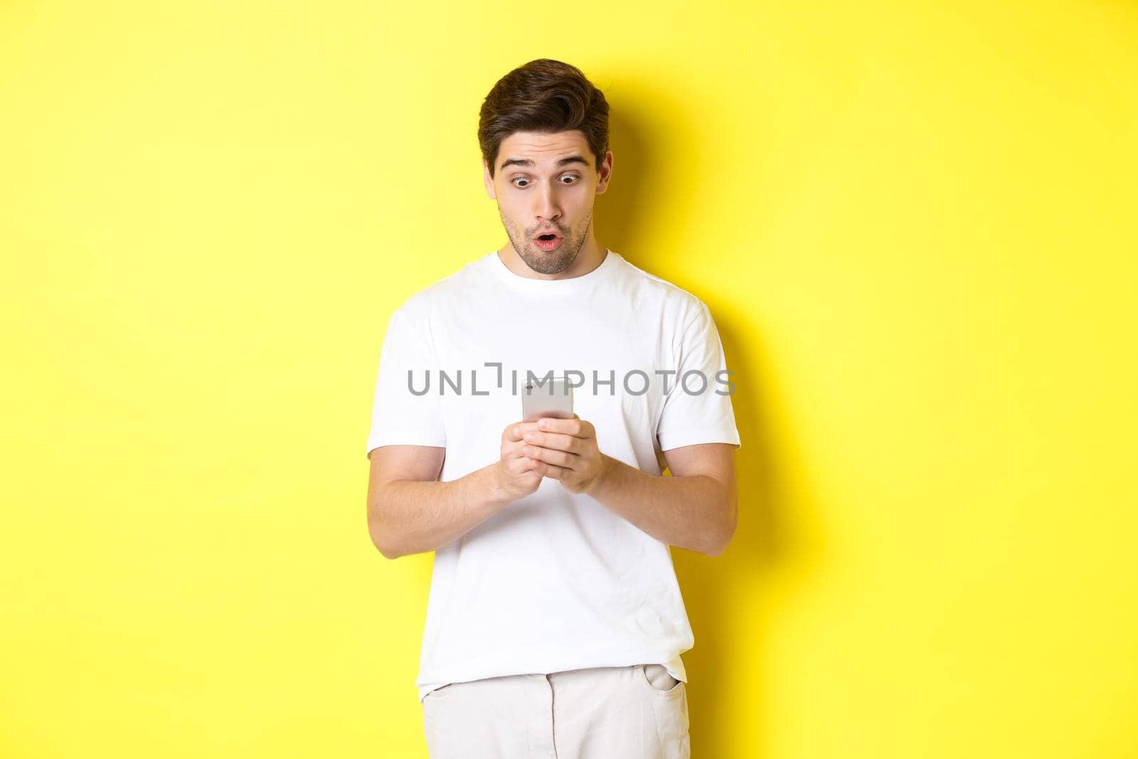 Man looking surprised in smartphone, reading message on cell phone, standing in white outfit against yellow background by Benzoix