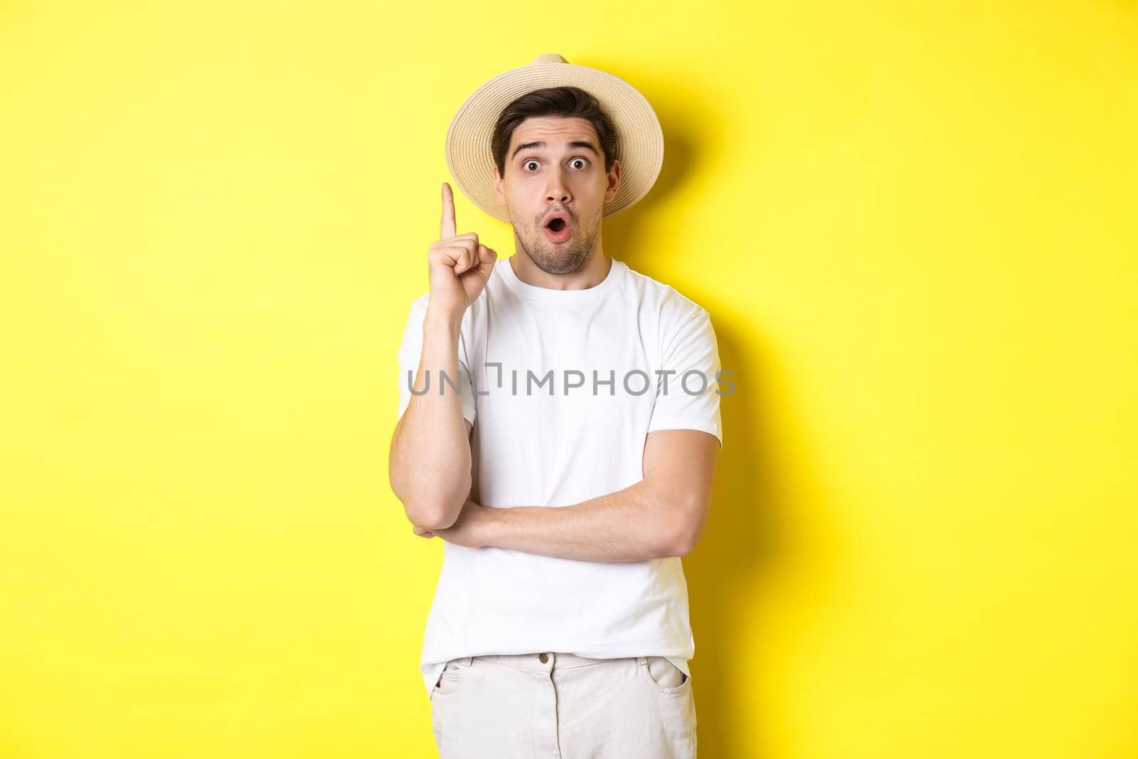 Portrait of young man in straw hat having an idea, raising finger eureka sign, making suggestion, standing over yellow background by Benzoix
