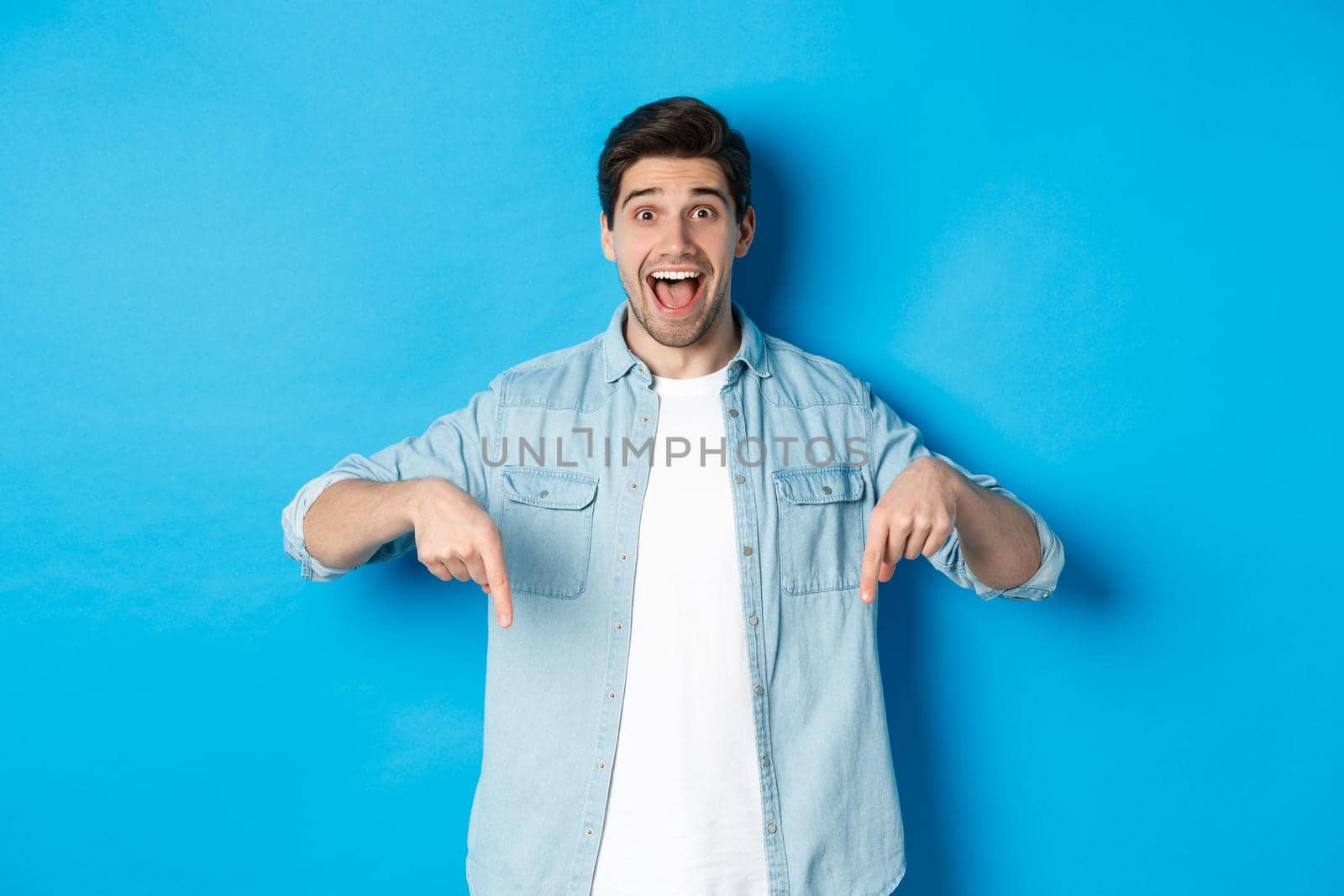 Image of excited handsome man pointing fingers down, making an announcement, standing against blue background.
