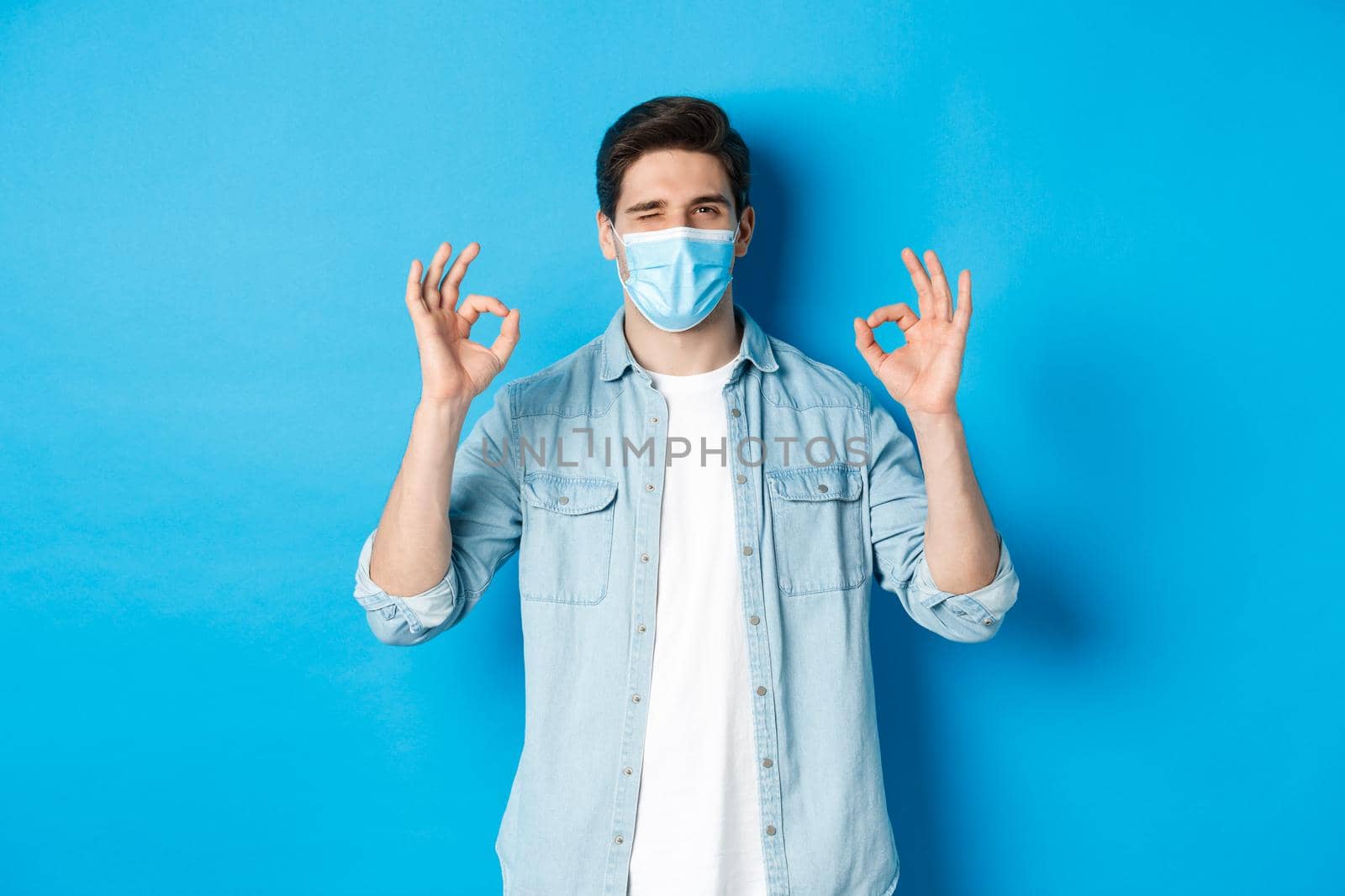 Concept of coronavirus, quarantine and social distancing. Cheeky man in medical mask winking, showing okay signs, assure or guarantee something, like and approve.