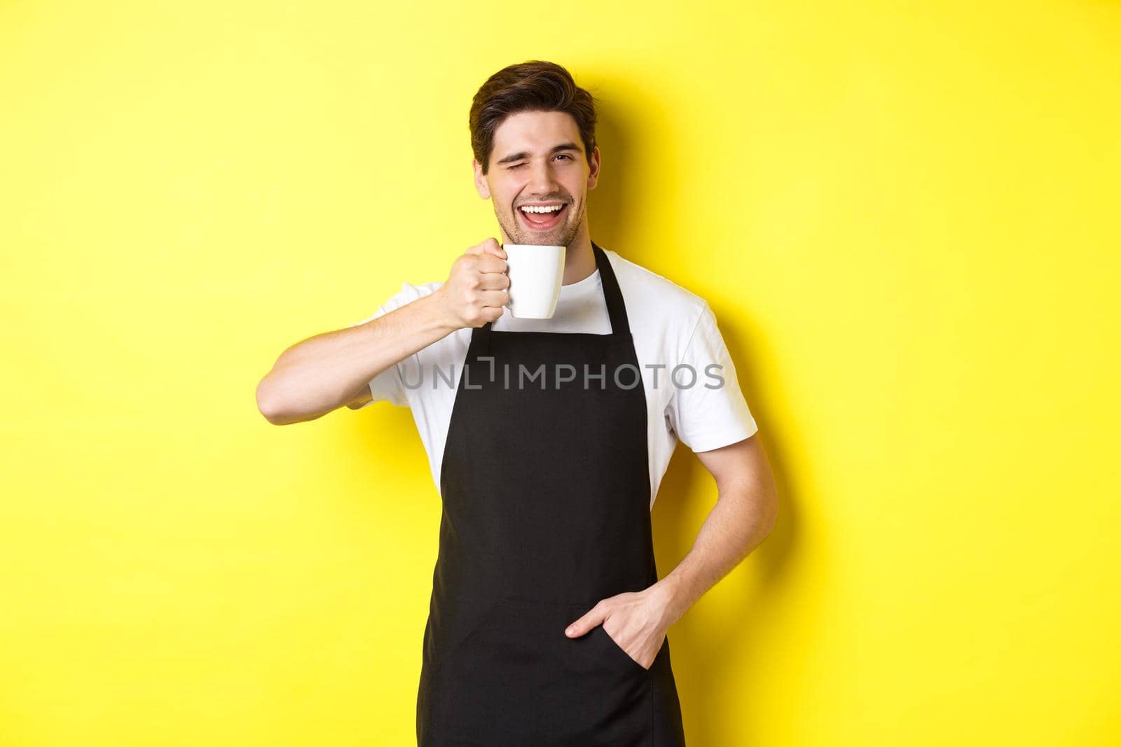 Handsome barista drinking cup of coffee and winking, inviting to cafe, standing over yellow background.