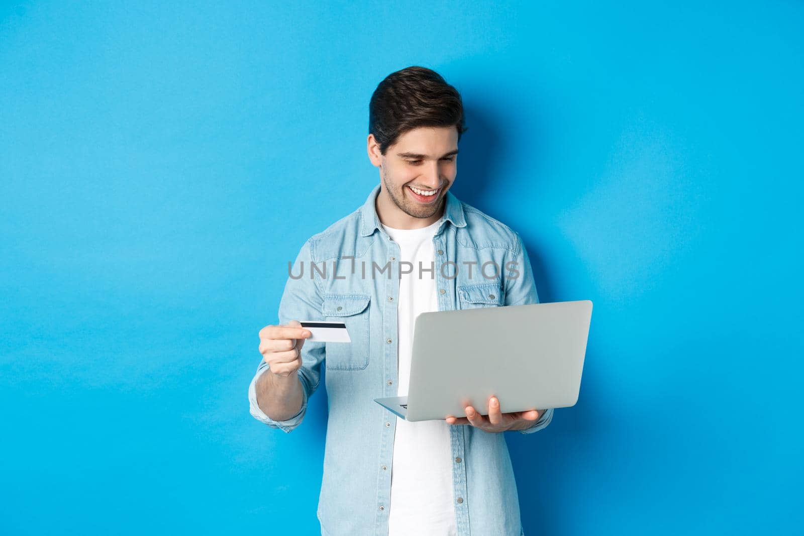 Young smiling man buying in internet, holding credit card and paying for purchase with laptop, standing over blue background.