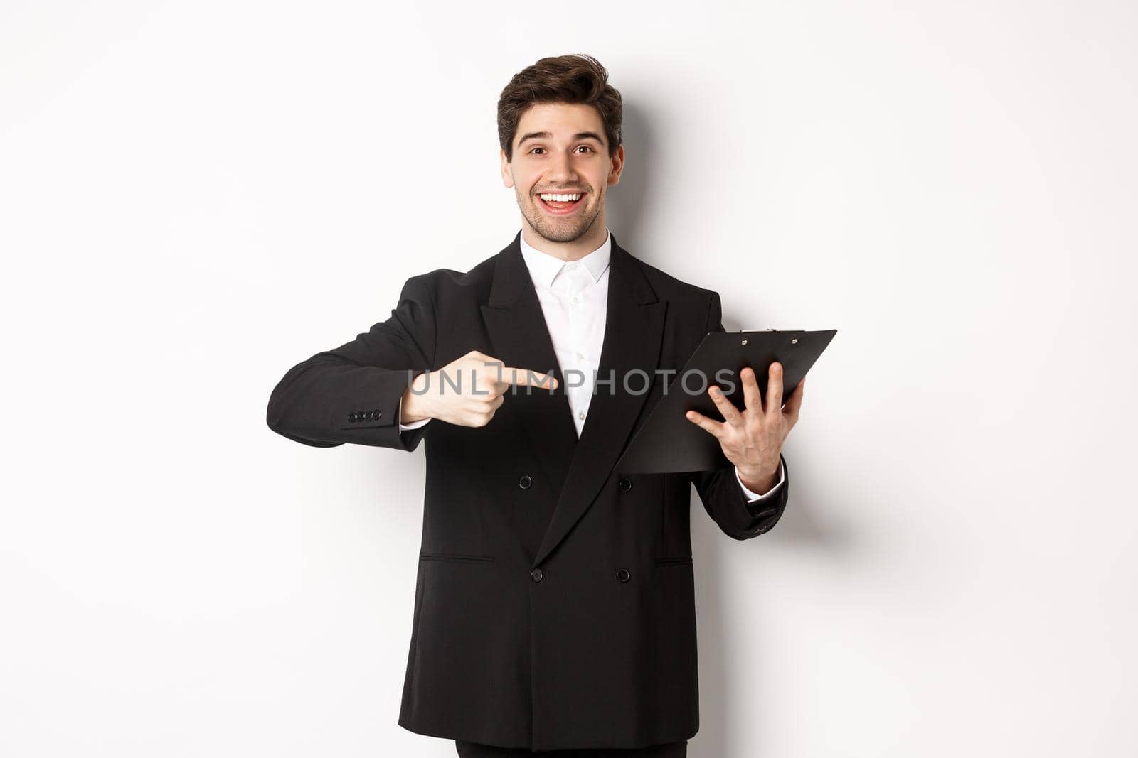 Image of handsome smiling businessman in black suit, pointing finger at clipboard with documents, standing against white background by Benzoix