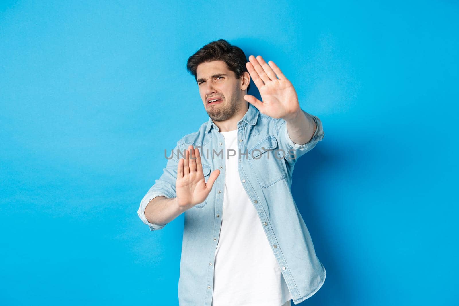 Disgusted guy saying no, refusing and looking away from something awful, cringe while standing against blue background.