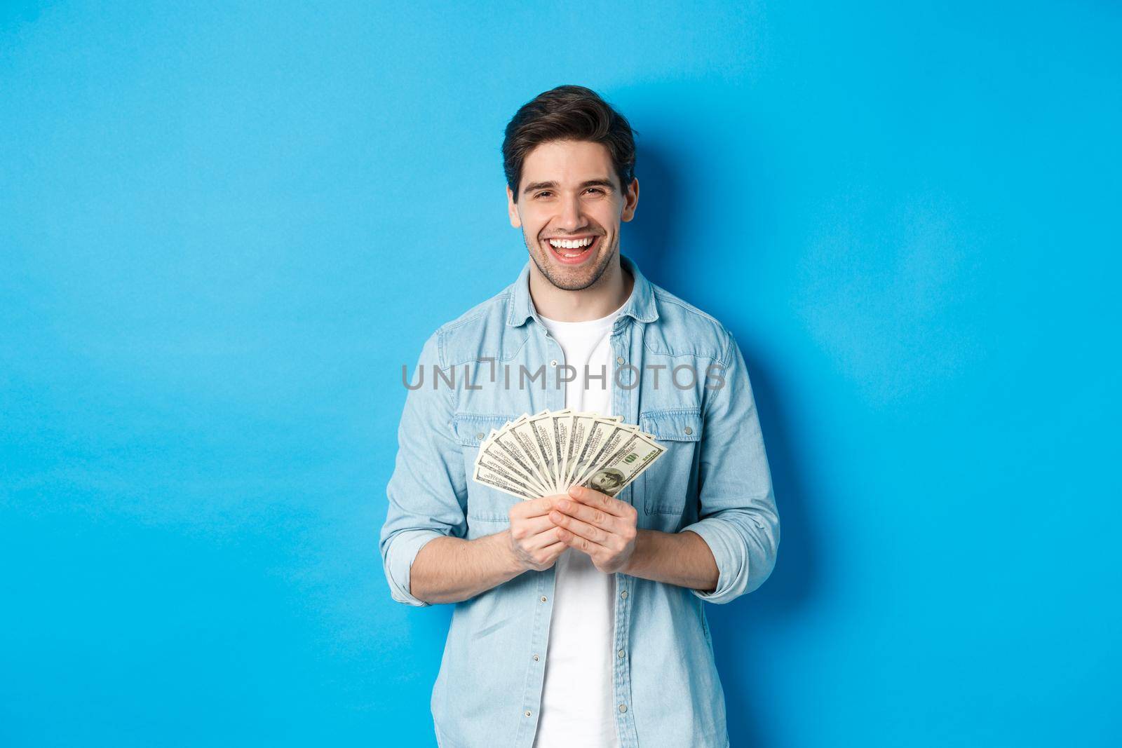 Happy successful man smiling pleased, holding money, standing over blue background.