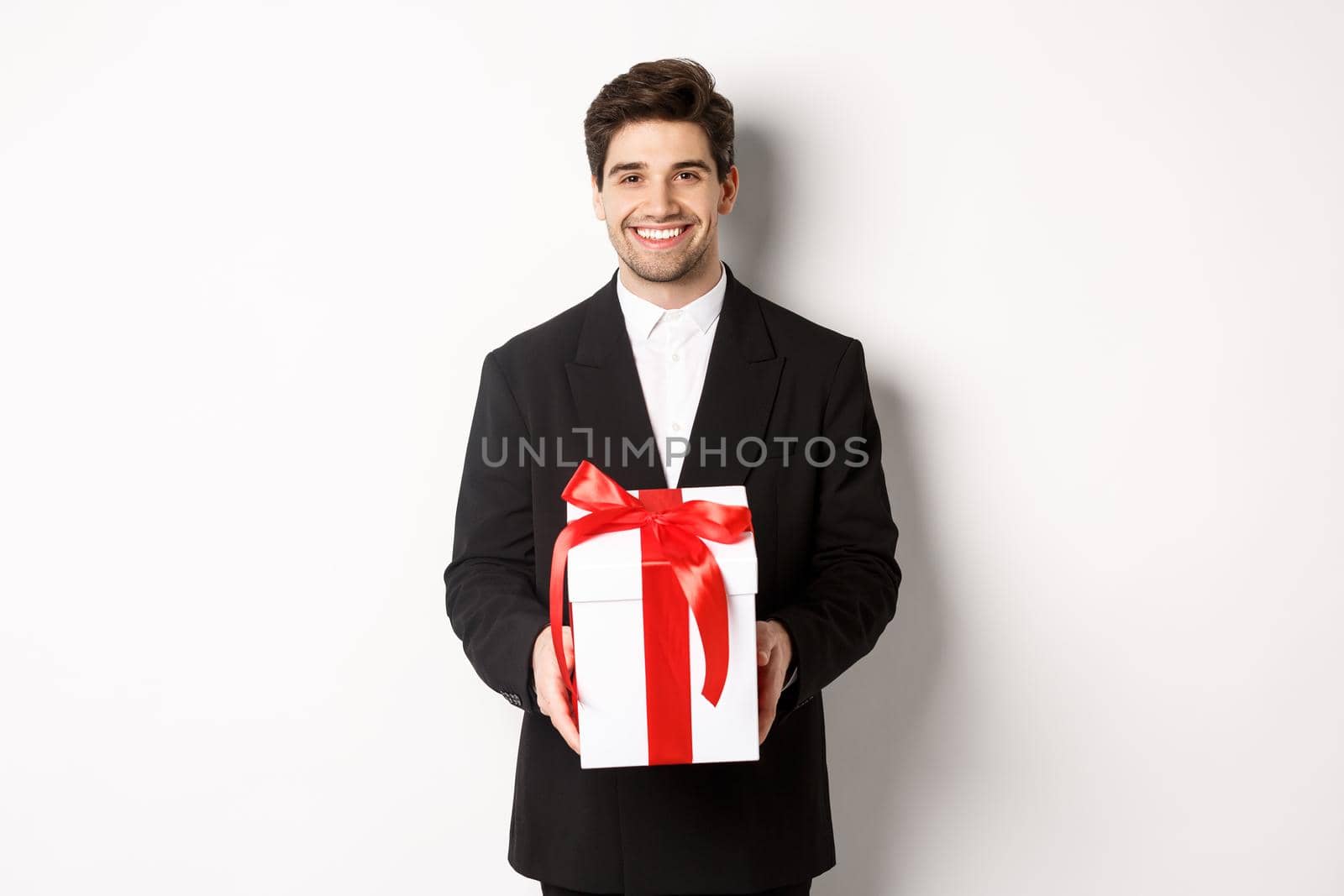 Concept of christmas holidays, celebration and lifestyle. Handsome man in black suit, have romantic present, holding gift in a box and smiling, standing against white background.