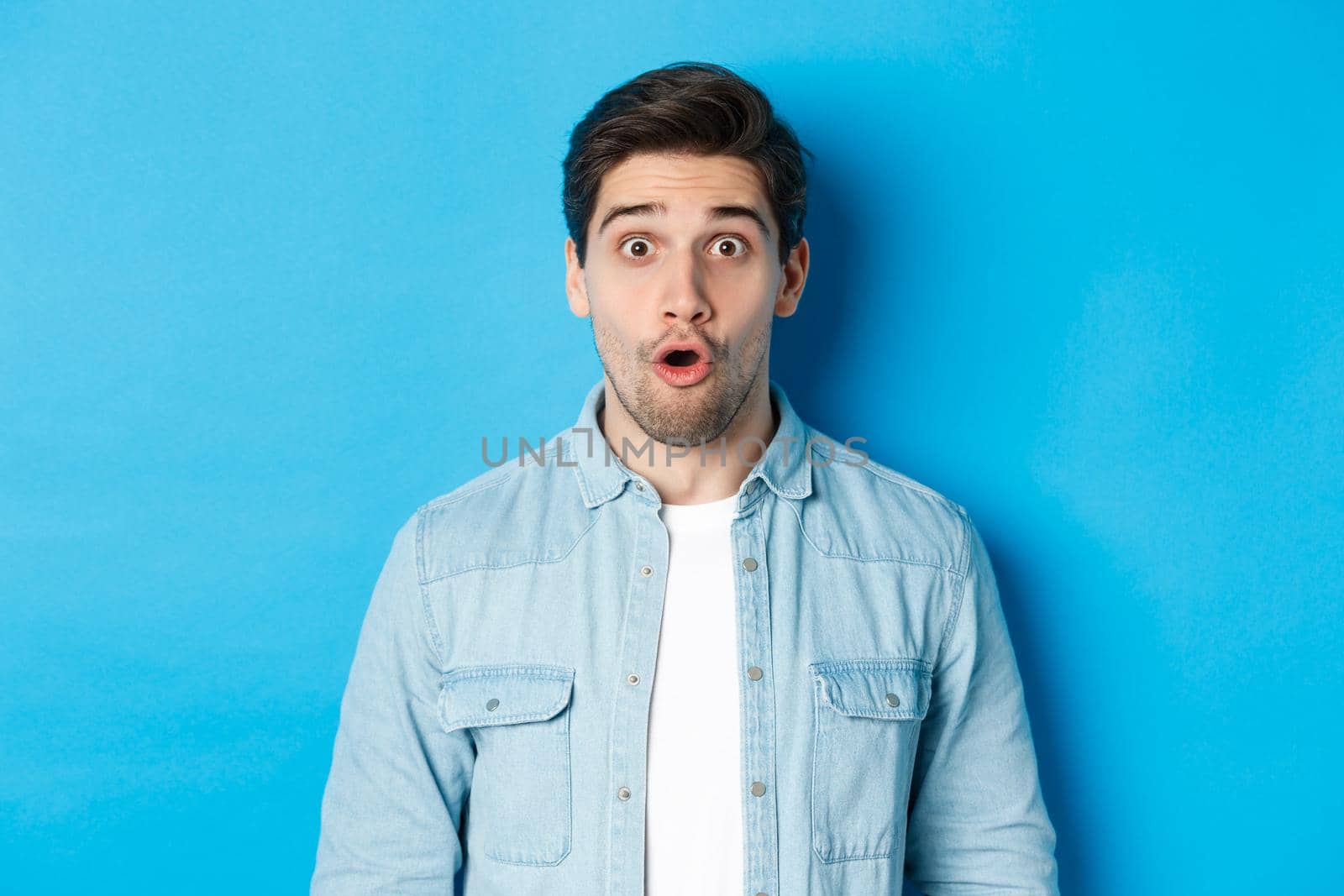 Close-up of handsome surprised man, open mouth and saying wow with amazement, hear announcement, standing over blue background.