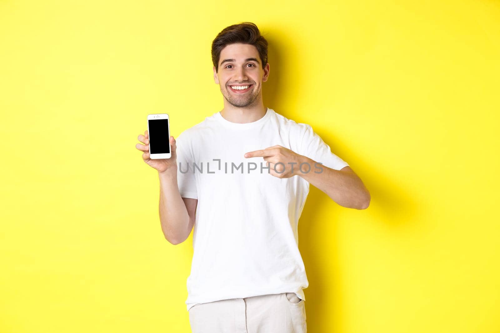 Image of attractive young man pointing finger at smartphone screen, showing an app, standing against yellow background.