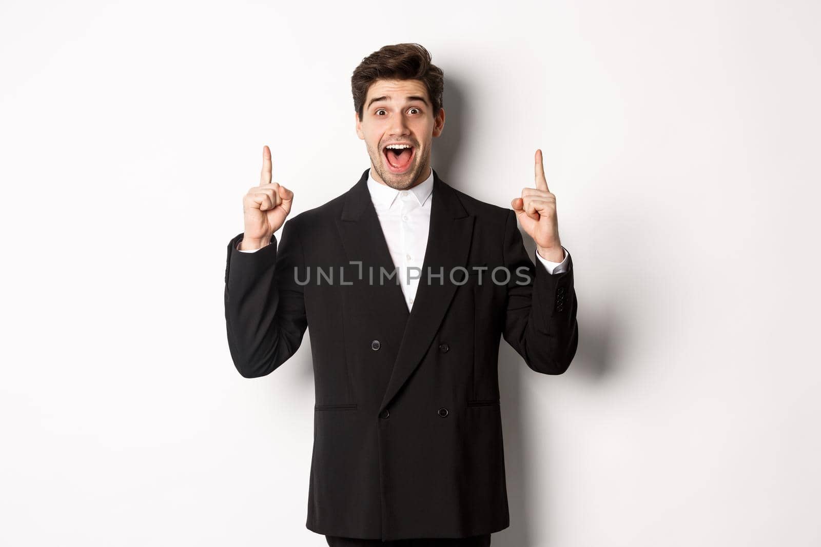 Image of handsome man in party suit, showing holidays promo, pointing fingers up and smiling amazed, standing over white background.