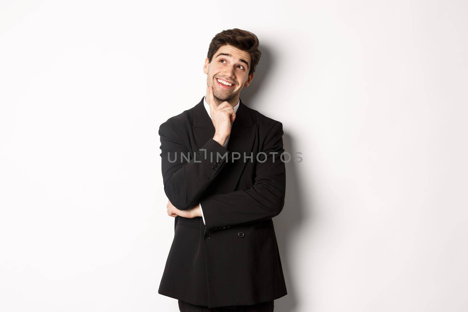 Portrait of thoughtful smiling man in stylish party suit, looking at upper left corner and thinking, imaging things, standing over white background.